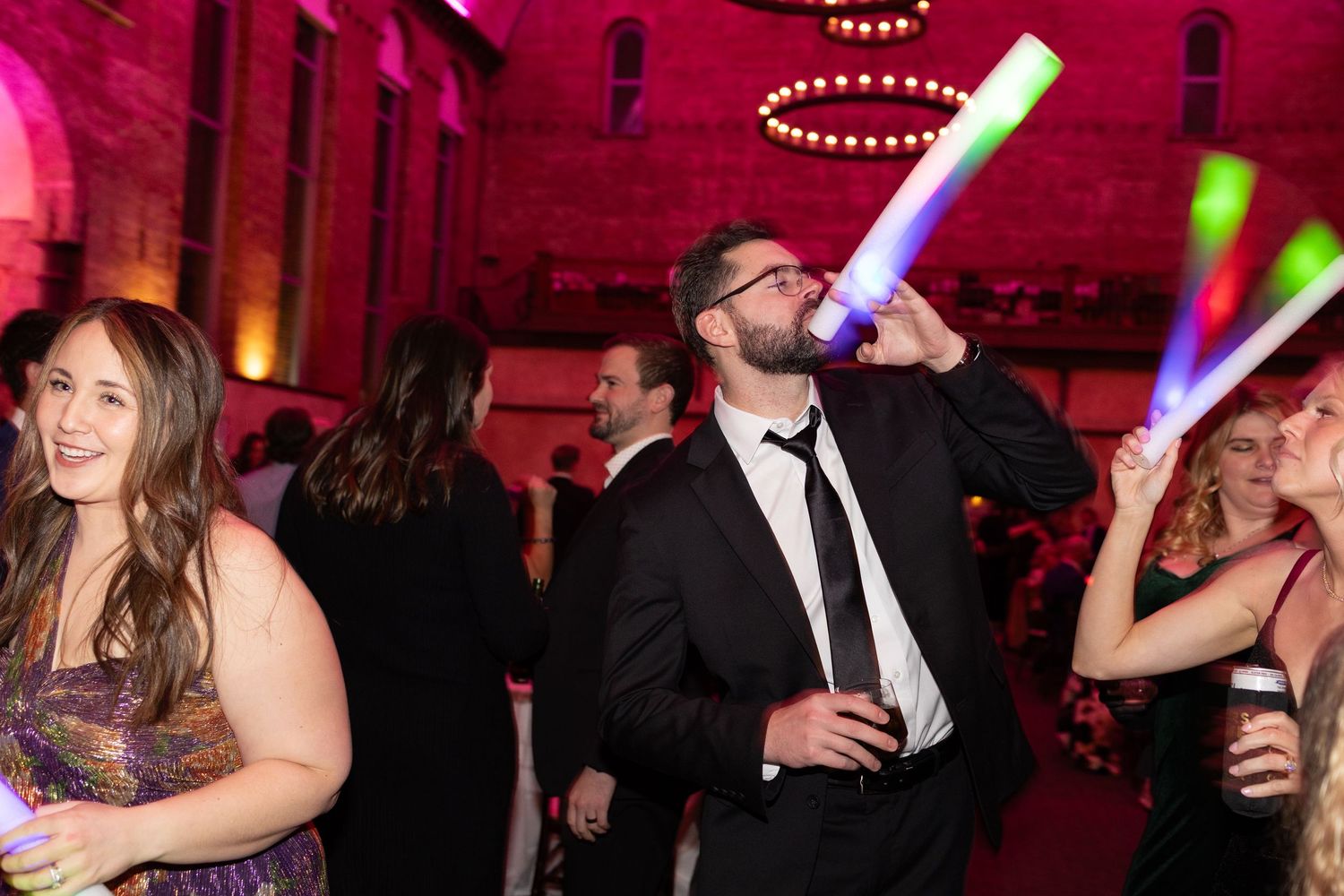 People dancing at a party with glowing foam sticks under pink lights at the historic Monastery Cincinatti wedding venue.