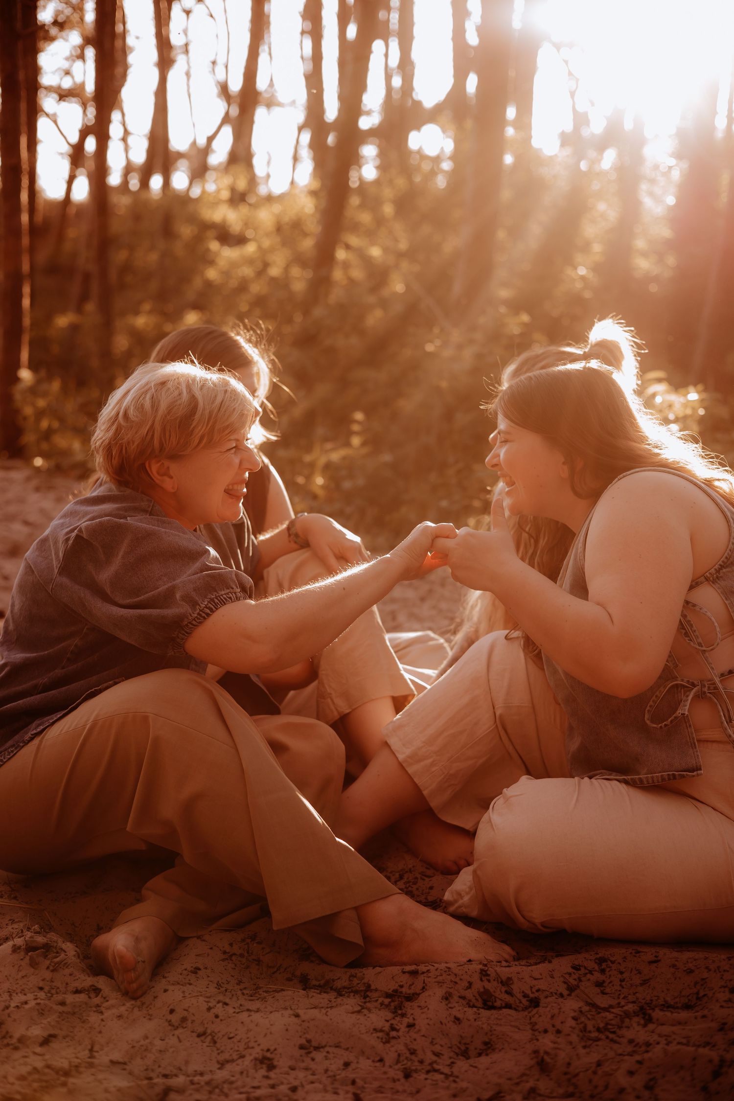 Two women sit on sandy ground in a sun-drenched forest, smiling and holding hands together.