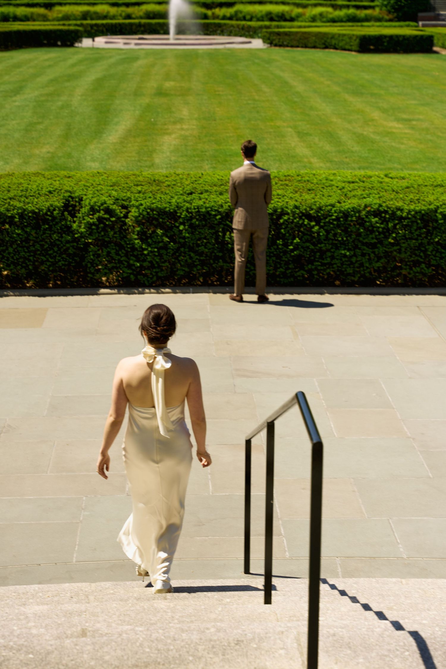 A couple in formal wedding attire embrace romantically in Central Park, NYC, with lush greenery and city skyline behind them.