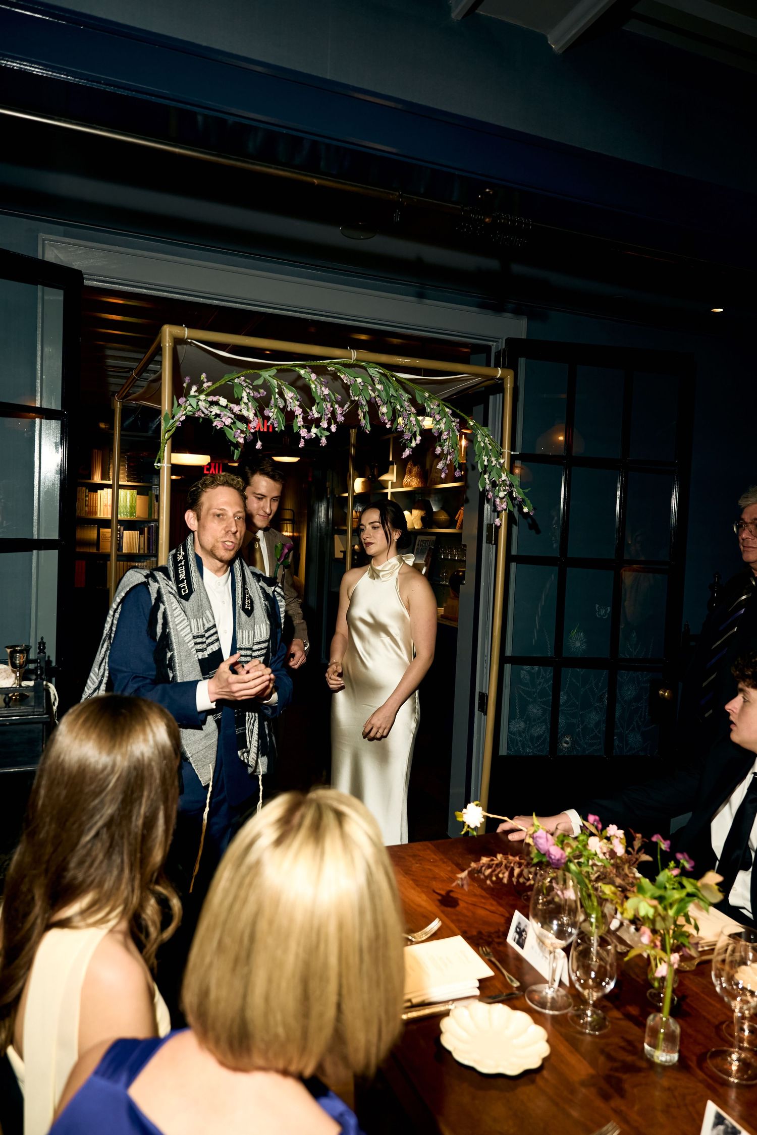 Bride and groom enter wedding reception under floral arch in elegant dark-toned venue with guests seated at wooden tables.