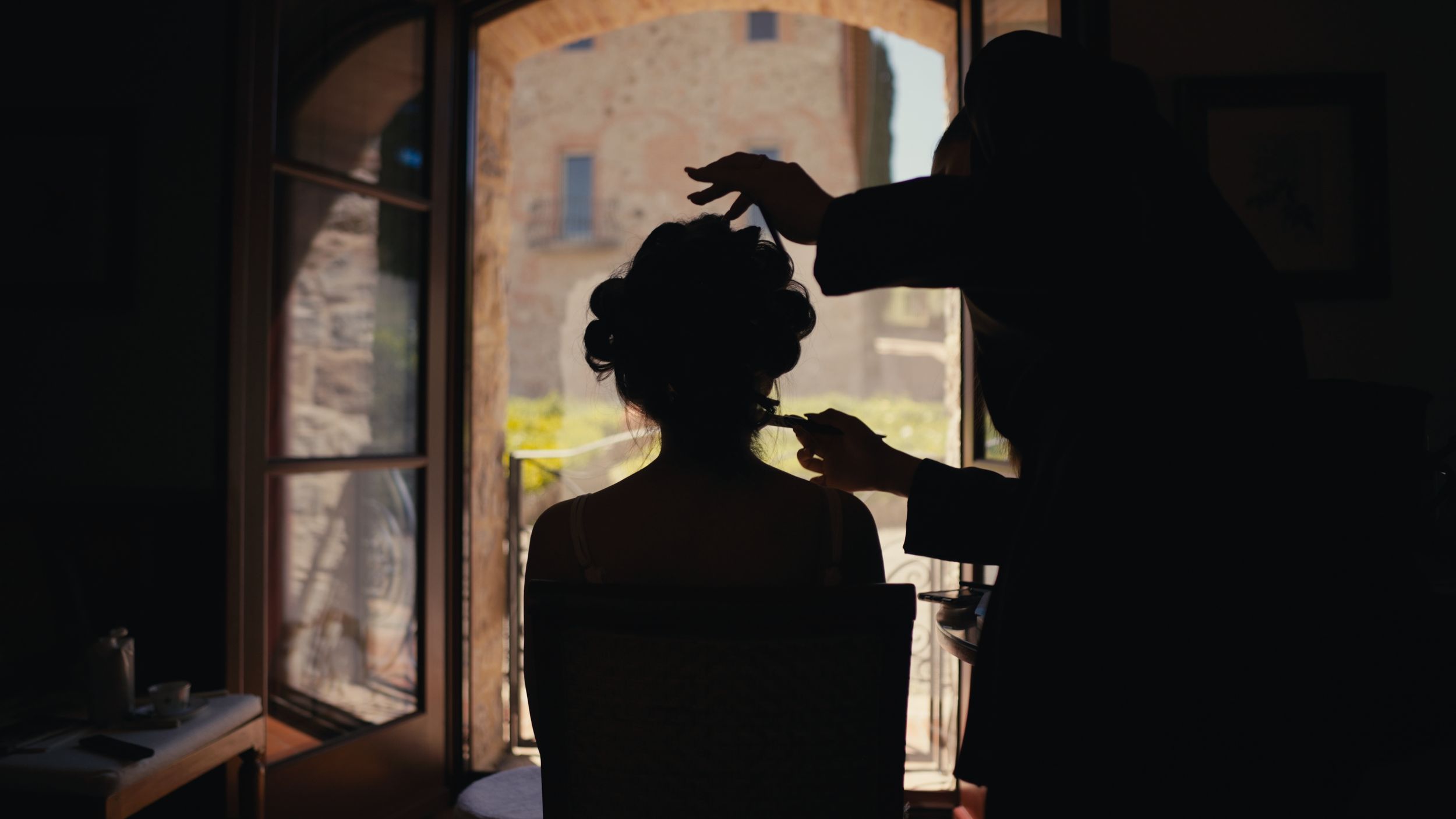 Silhouette of a bride getting her hair styled by a hairdresser in front of a bright window.