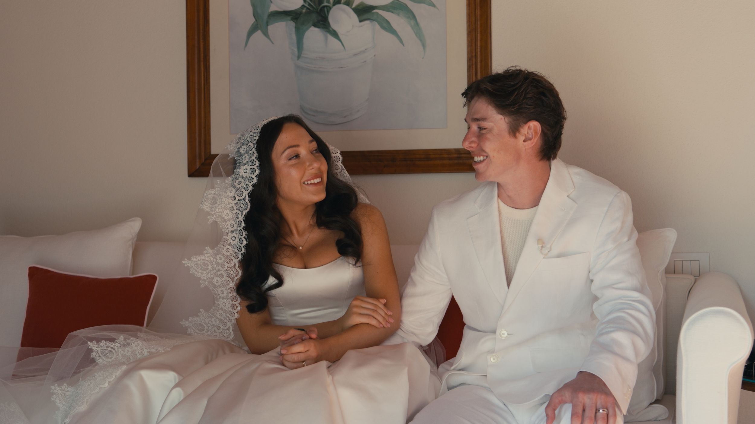 A bride in a white strapless gown and groom in a white suit sit together smiling on a white sofa.