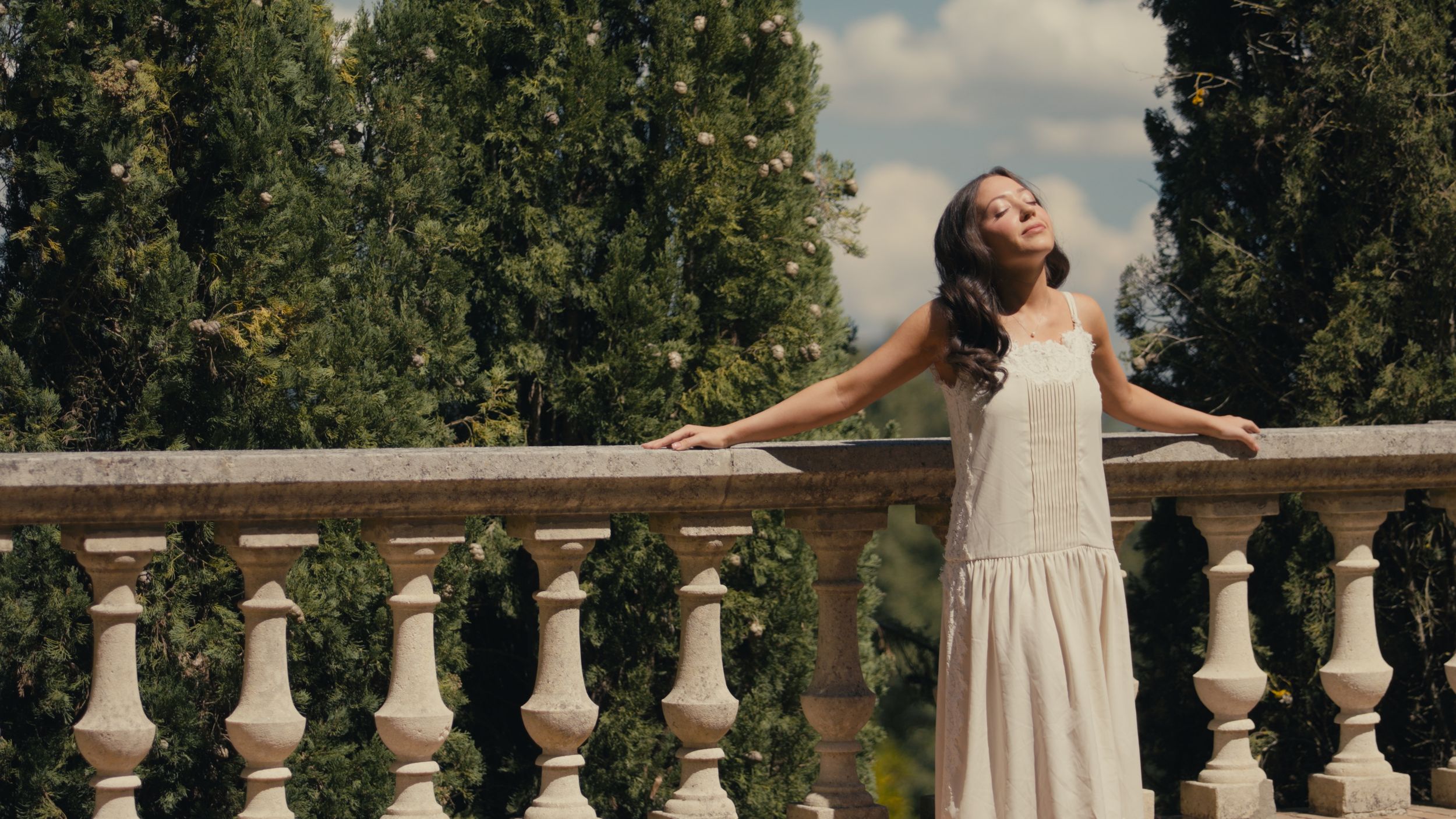 A woman in a white dress poses gracefully on a stone balcony surrounded by lush green trees.