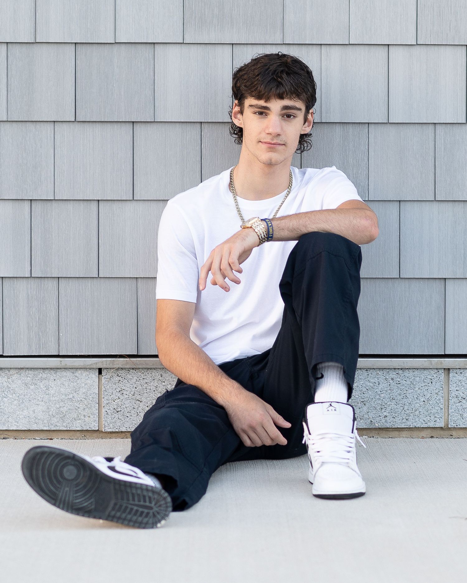 Young man in white t-shirt, dark pants, and white Air Jordans sitting casually against a gray shingle wall.