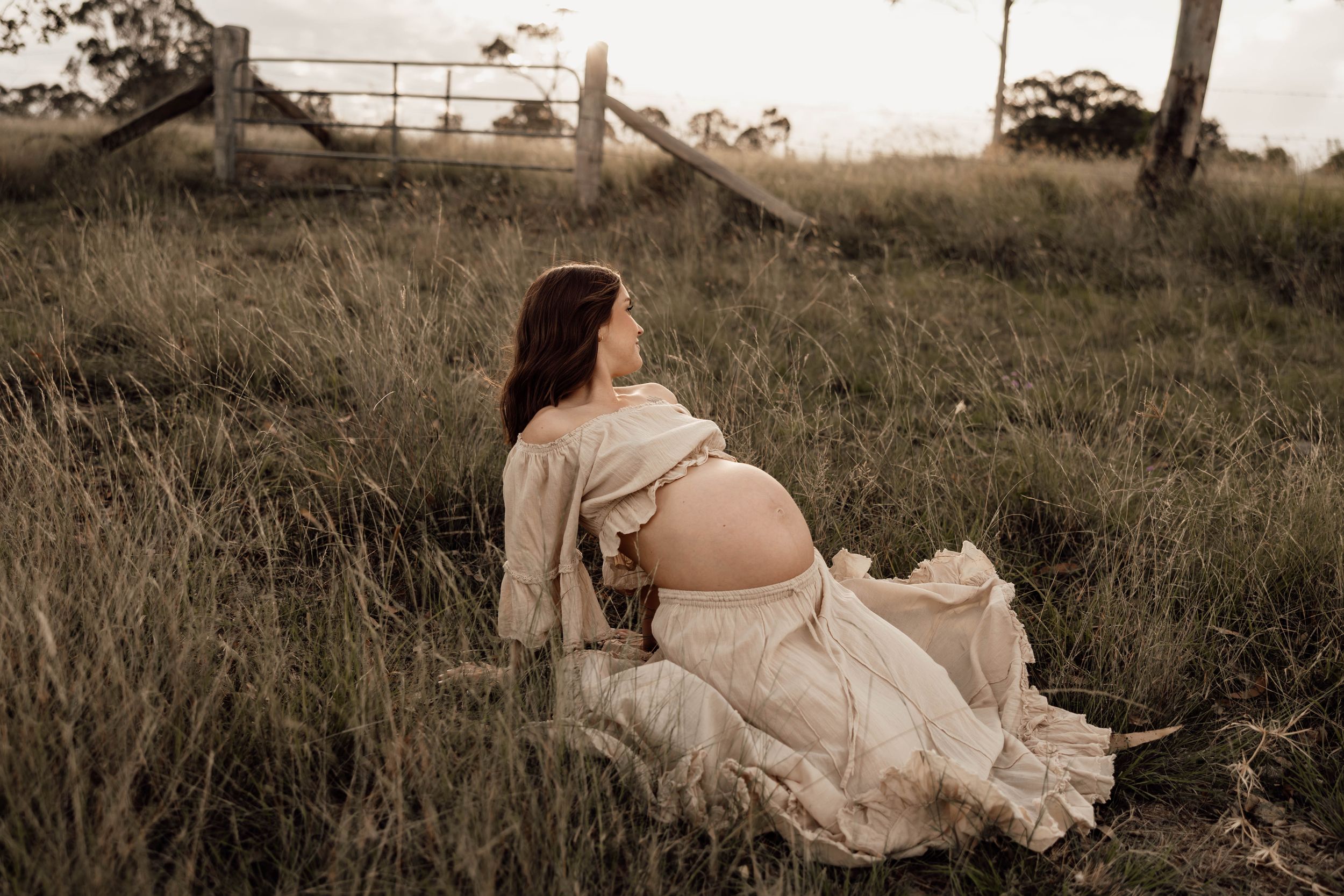 Pregnant woman in cream dress sitting in tall grass at sunset near rustic wooden fence.