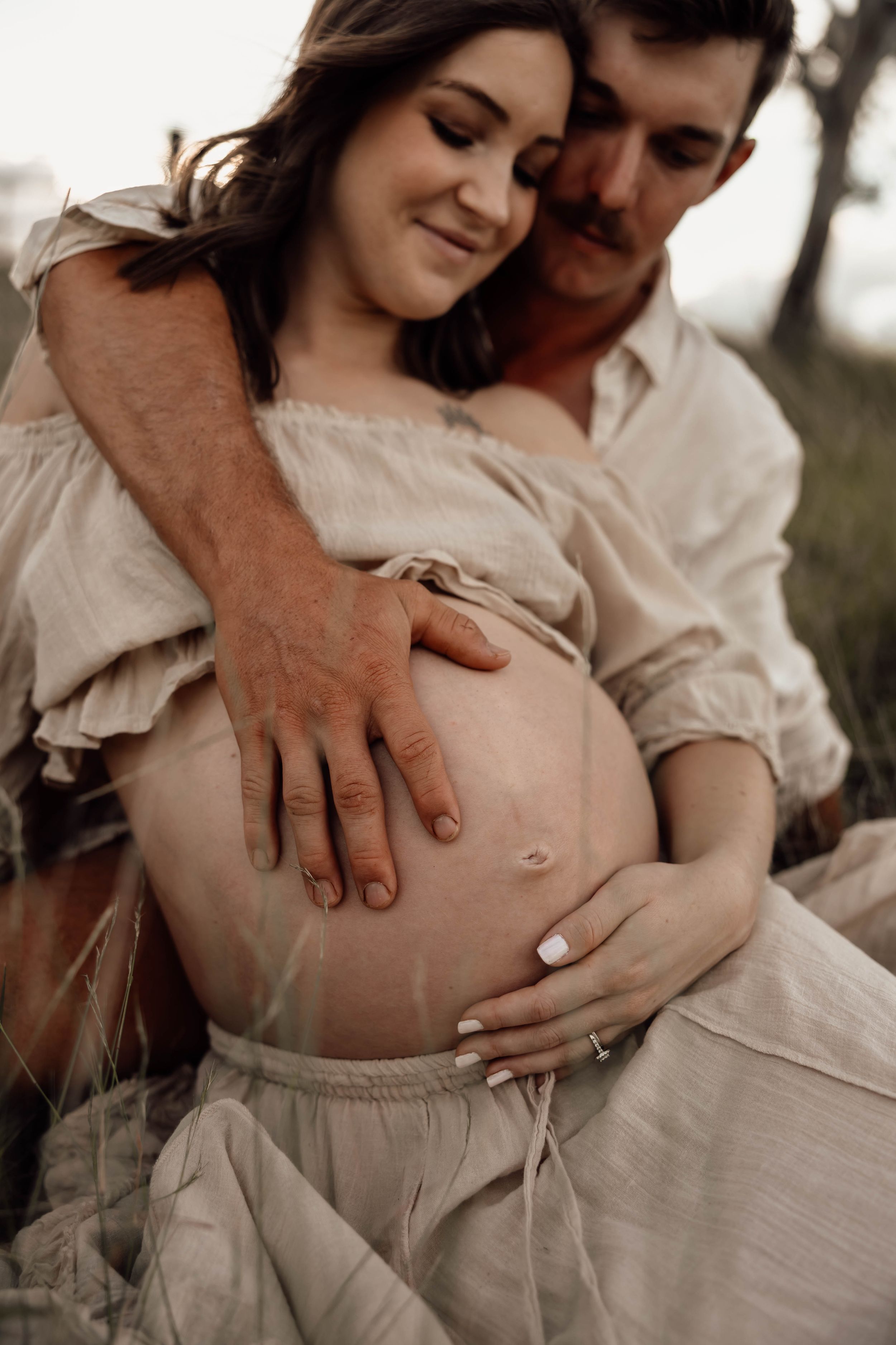 Couple tenderly holding pregnant belly together while sitting in grassy field.