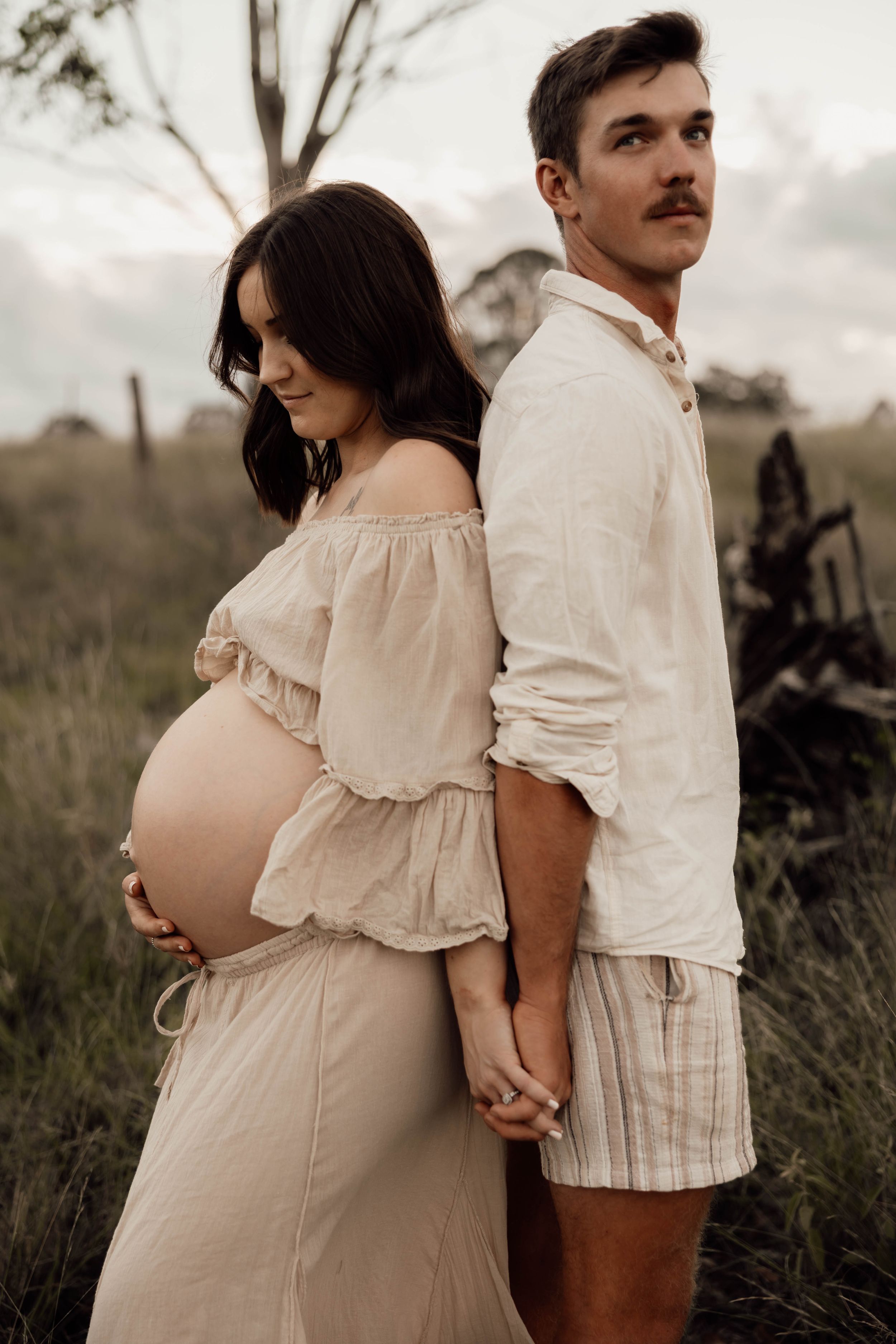 Pregnant couple standing back-to-back in a field, woman in beige off-shoulder dress cradling baby bump, man in white linen outfit.