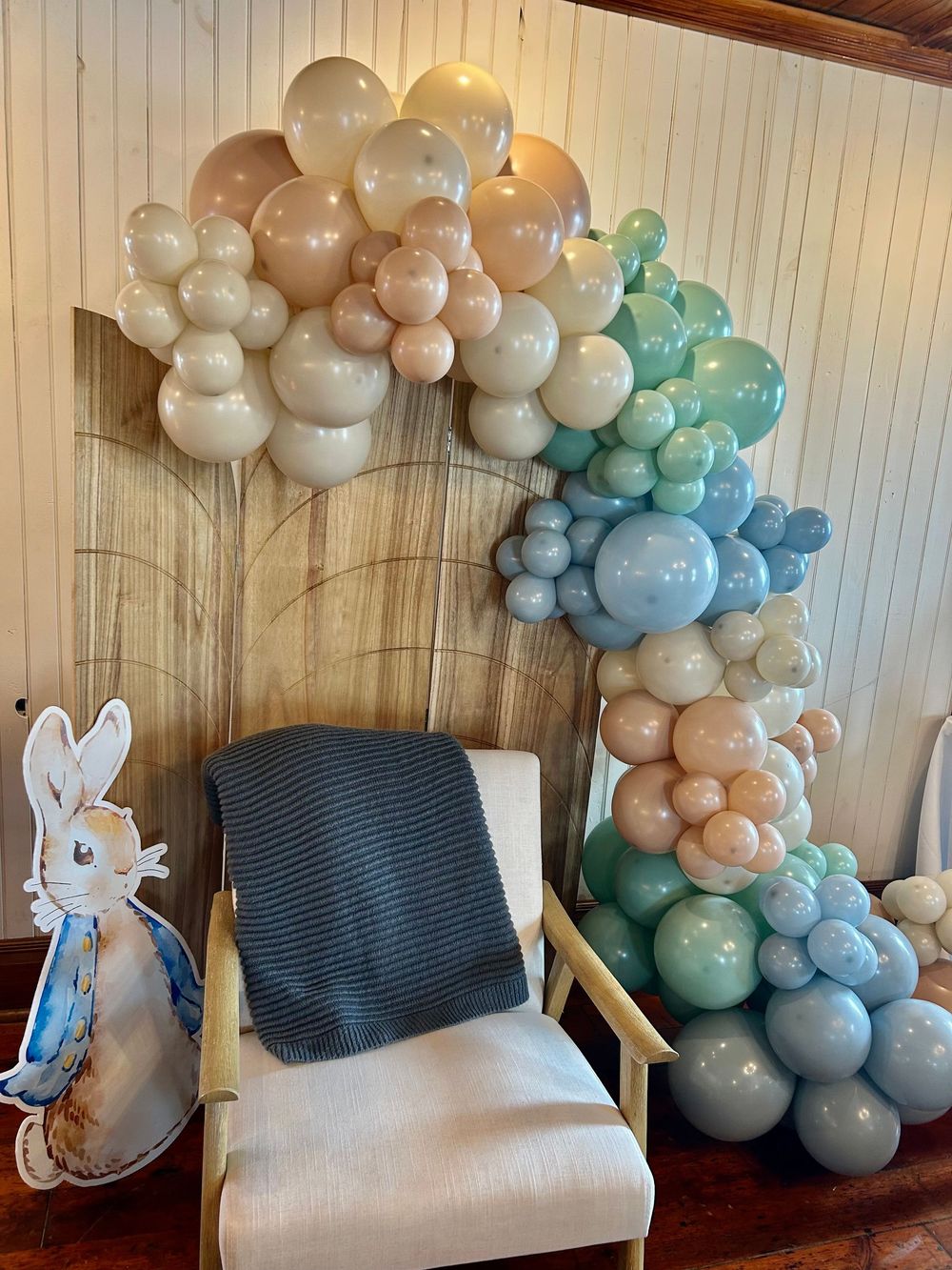 Pastel balloon garland arch in peach, sage, and blue tones framing a white chair with gray blanket and Peter Rabbit cutout.