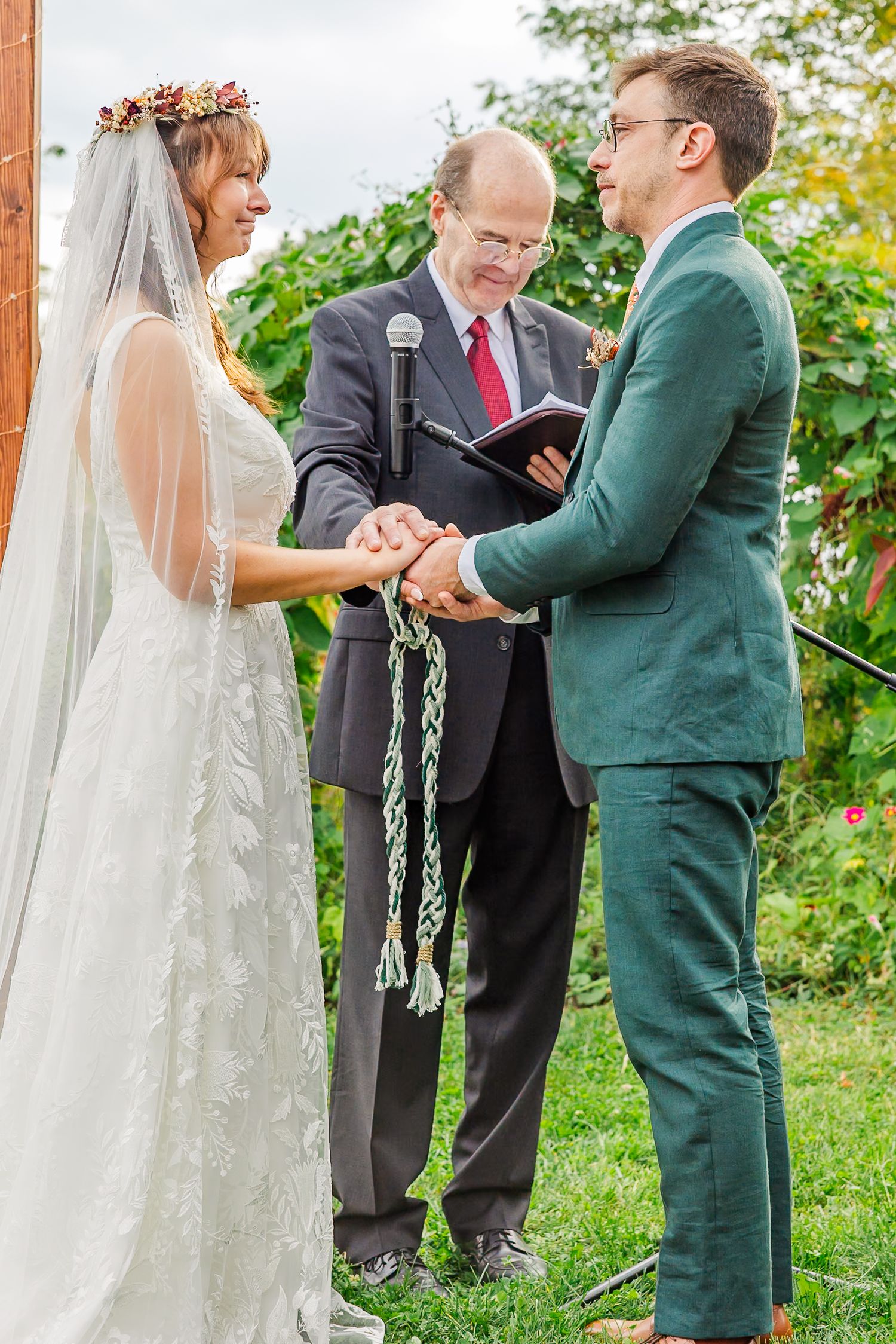Bride and groom hold hands during outdoor wedding handfasting ceremony while officiant reads from book.