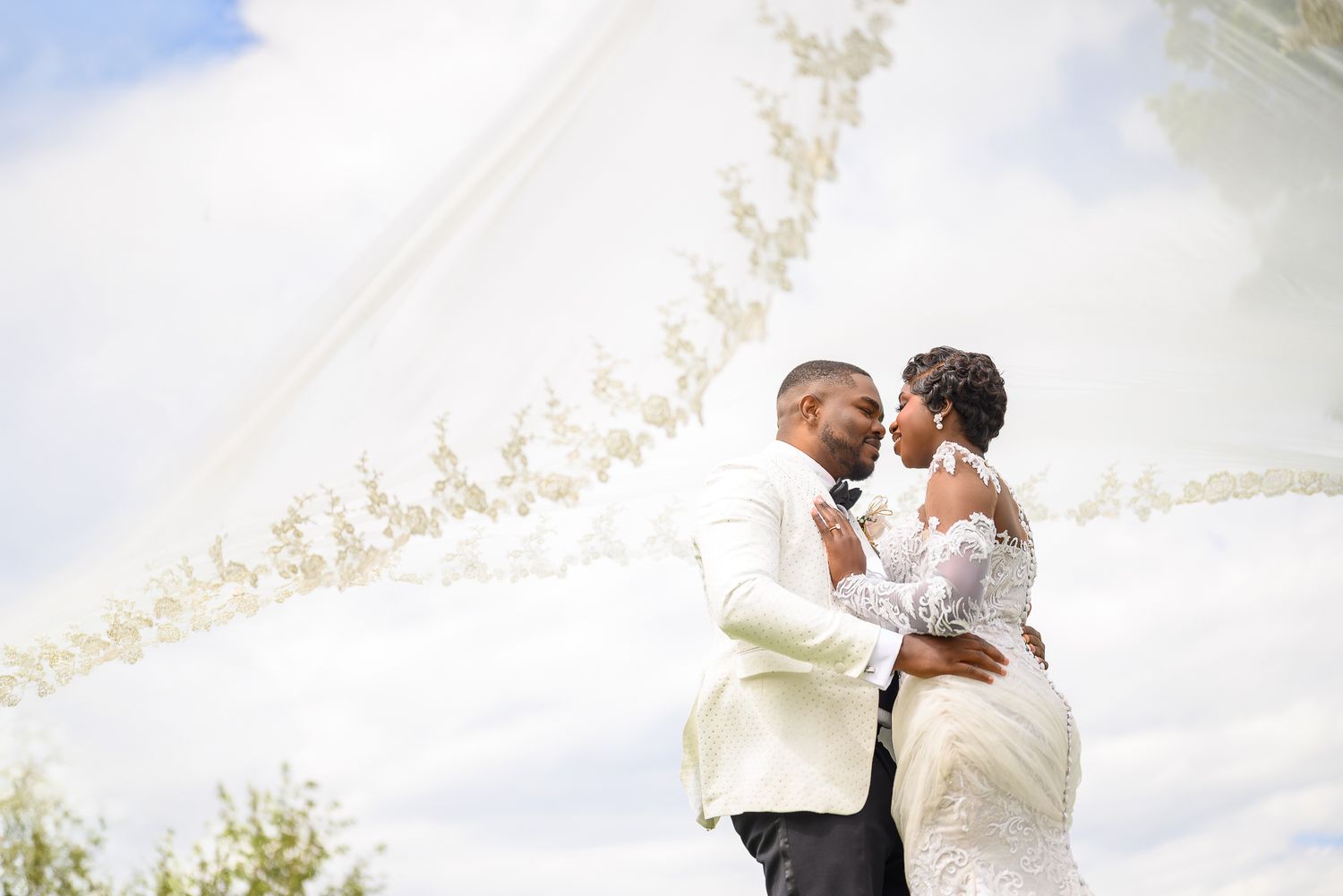 A groom in a white suit lifts his bride in a lace gown outdoors, her veil billowing against a cloudy sky.