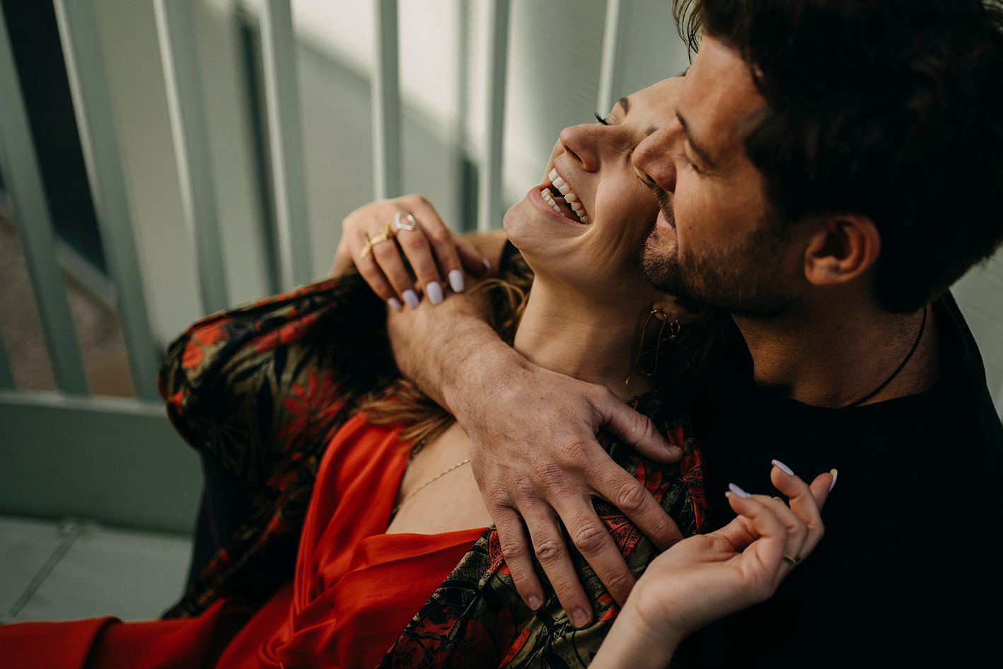 Couple sharing intimate moment, woman laughing as man nuzzles her cheek, wearing red floral dress.