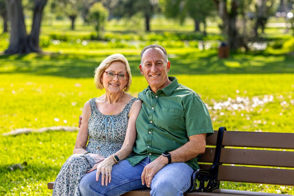 A couple sits together on a wooden bench in a sunlit park with blooming flowers in the background.