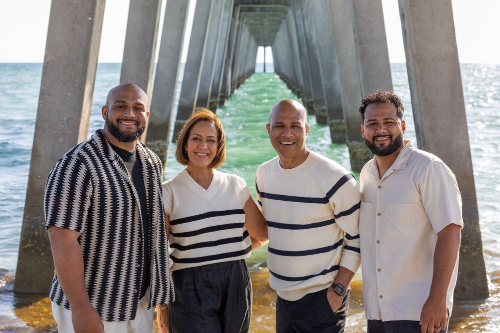 Four people smiling under a pier with ocean waves and perspective of columns in background.