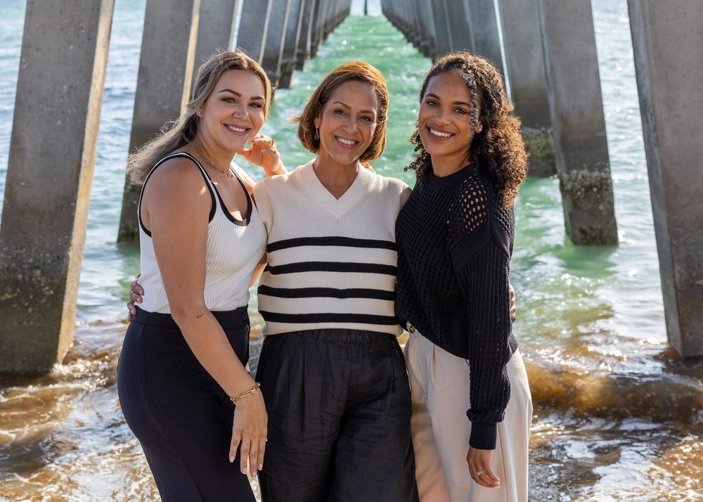 Three people pose together under a pier with ocean waves visible through the columns.