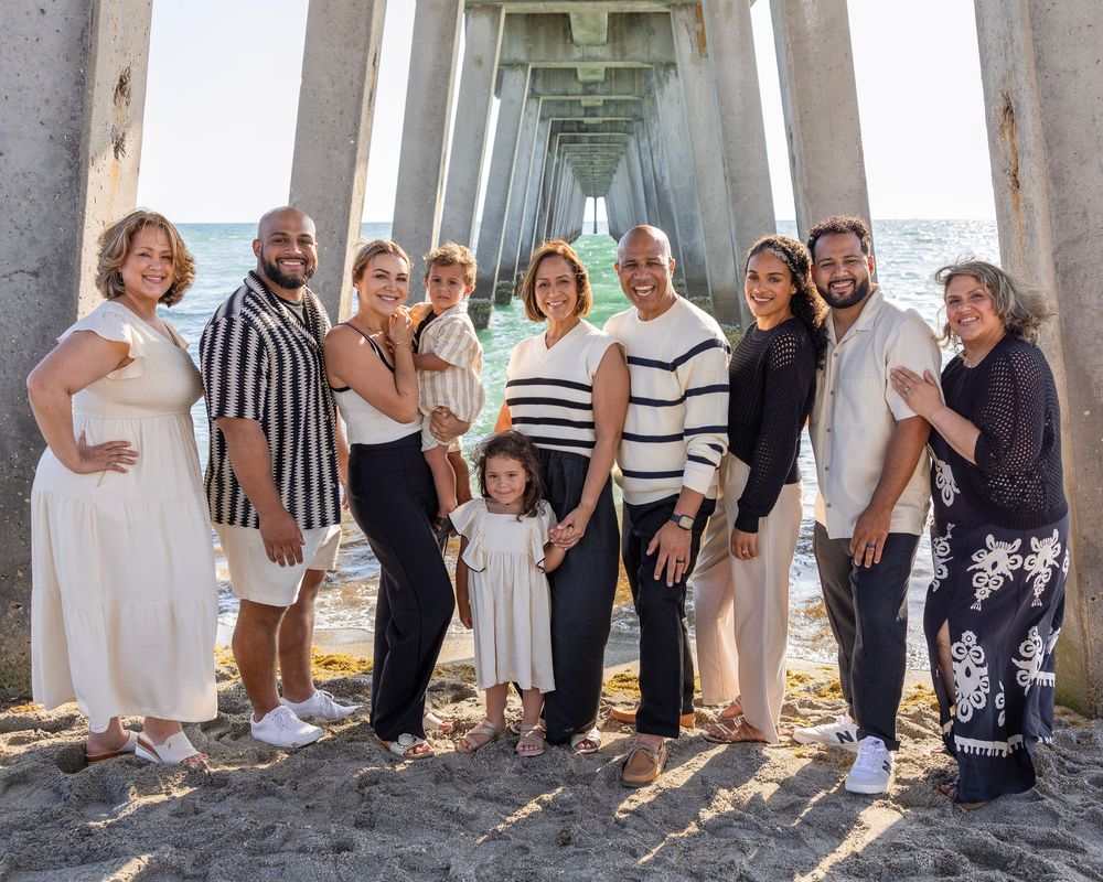 Extended family group photo at the beach under a pier wearing coordinated casual outfits.