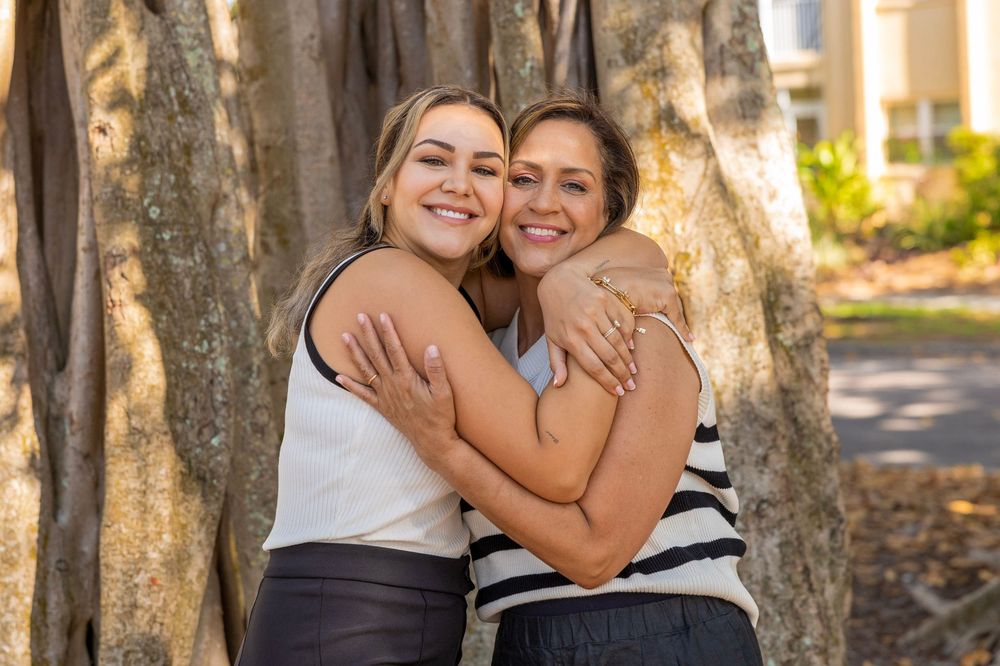 Two people share a warm embrace while laughing outdoors near a tree.