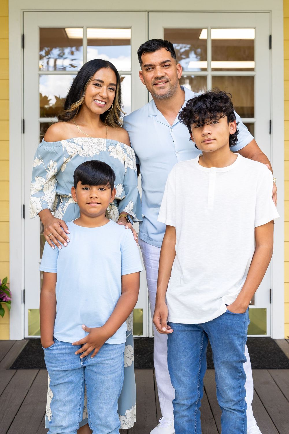 A family of four poses together on a front porch wearing coordinated blue and white casual outfits.