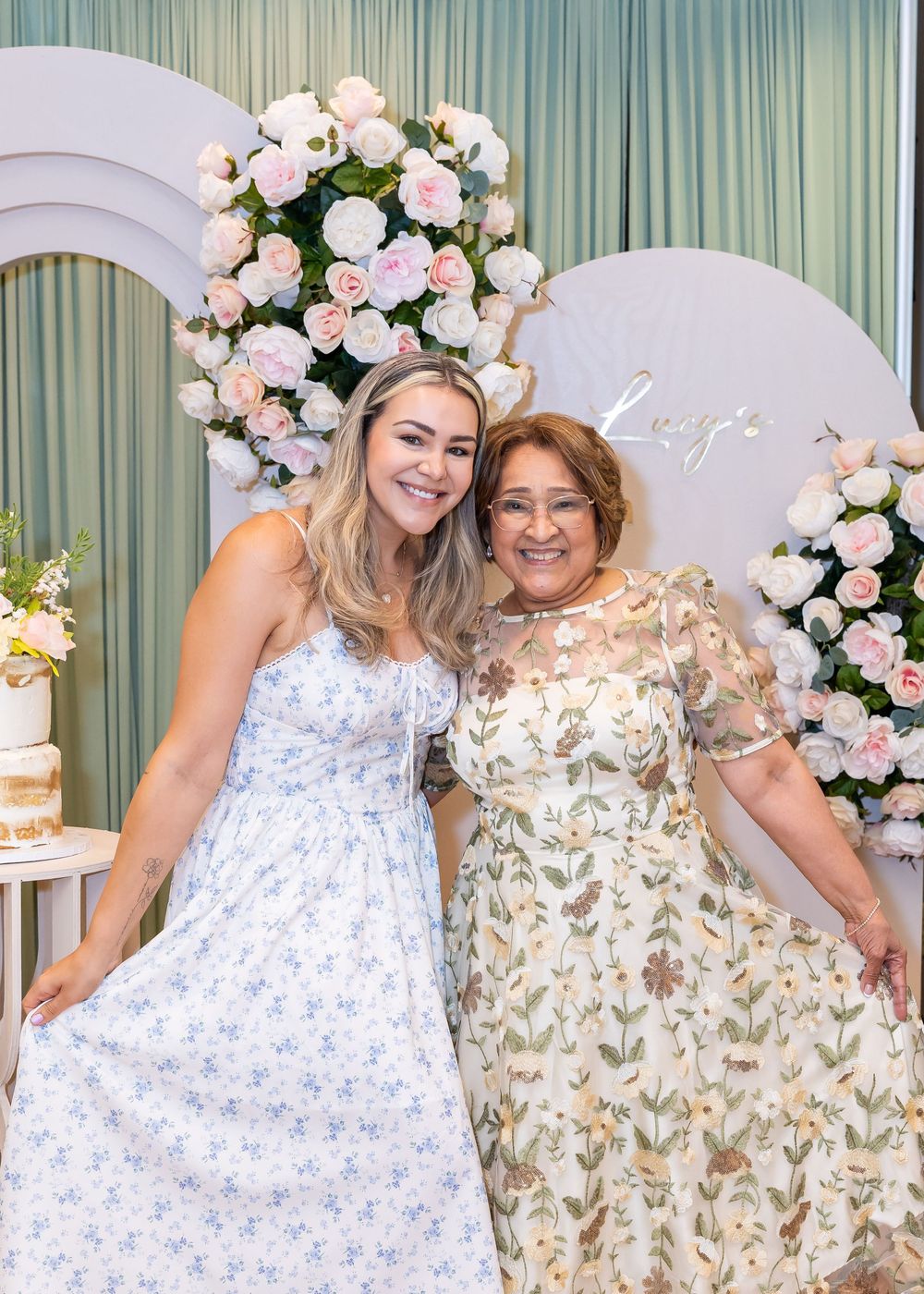 Two people in floral dresses pose together at an elegant event with pink roses and a decorative arch backdrop.