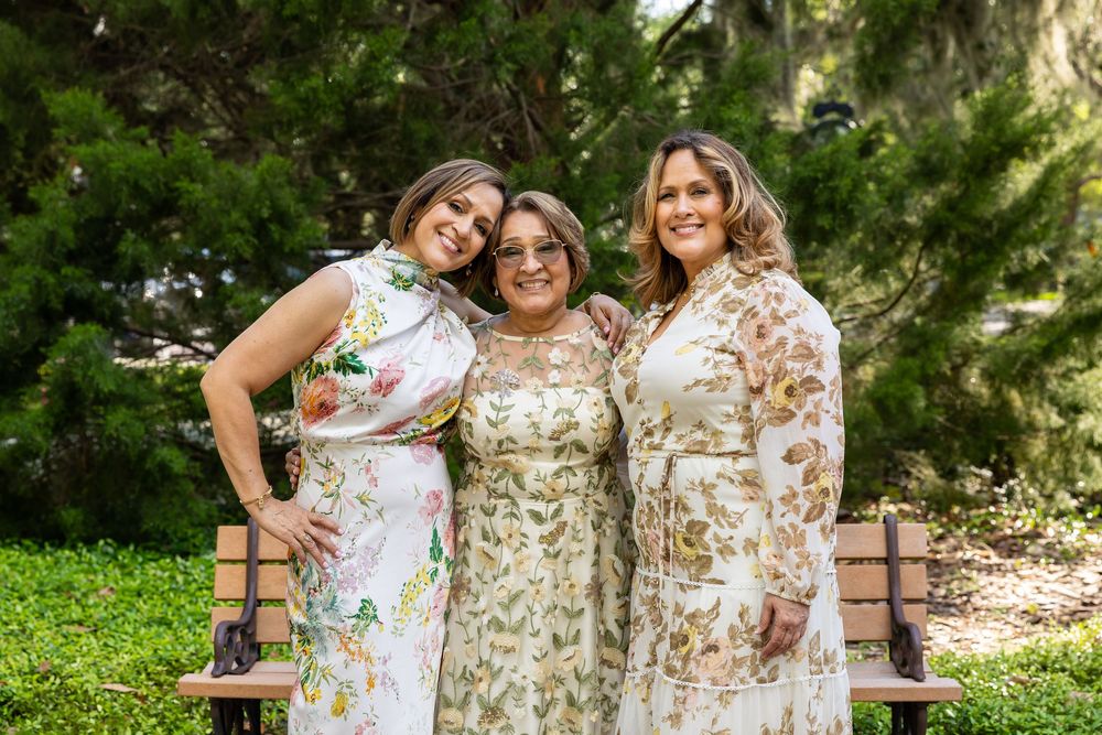 Three people wearing coordinated floral dresses pose together outdoors on a wooden bench.
