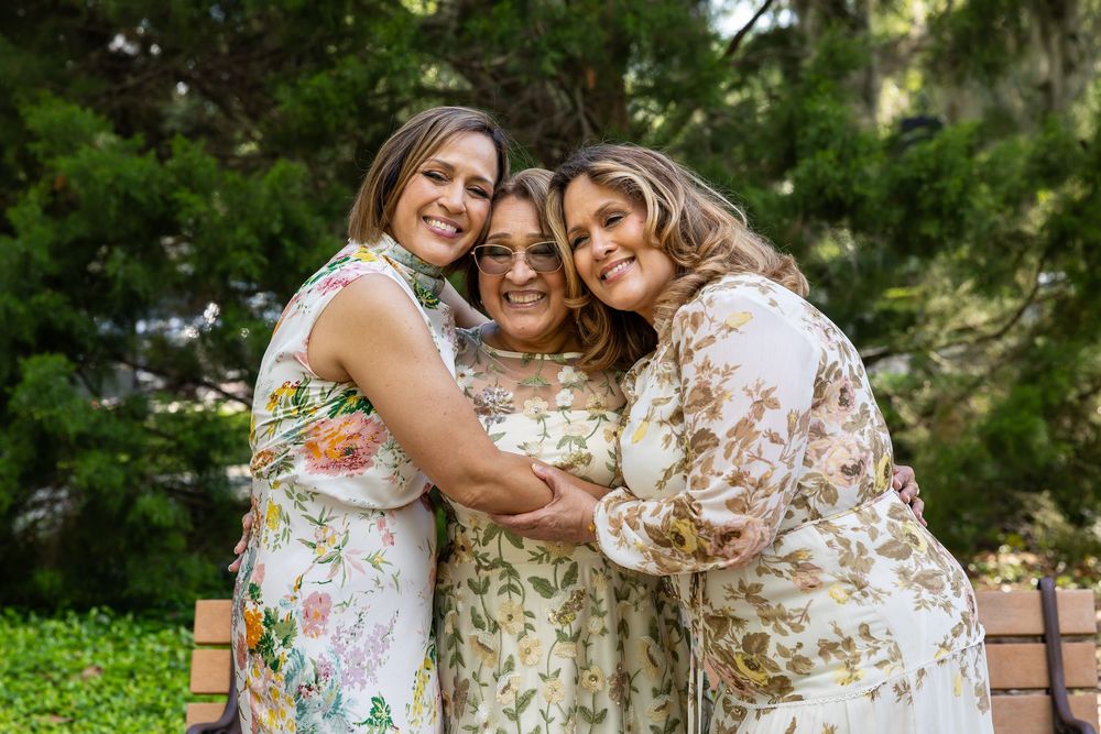 Three people embrace while wearing coordinated floral dresses in a garden setting.