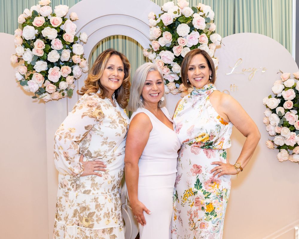 Three people in floral and white dresses pose against a pink rose arch backdrop at an elegant event.