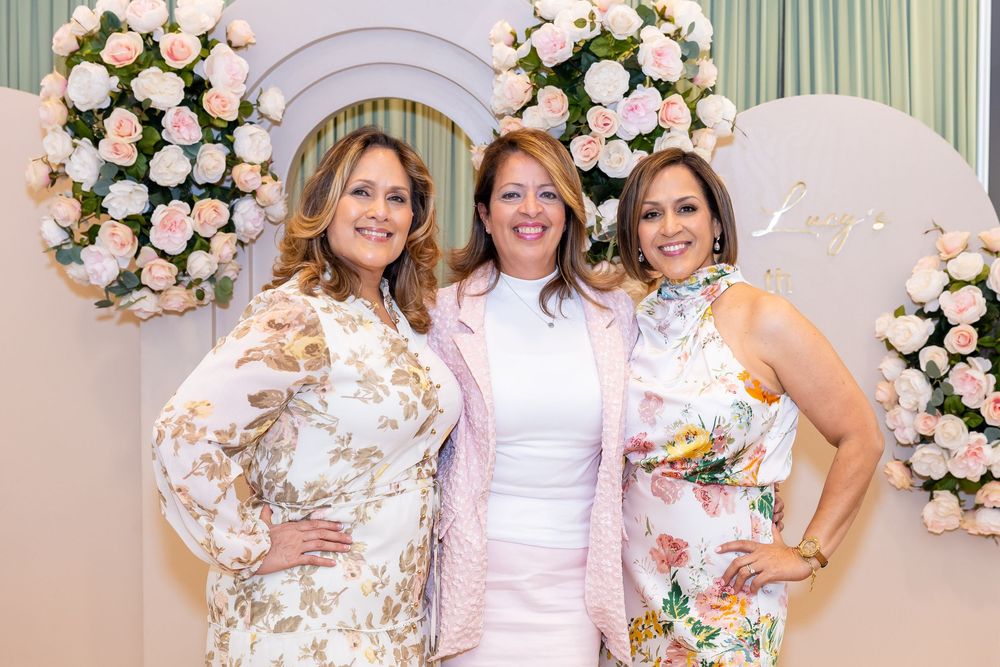 Three people pose in front of a floral arch wearing coordinated white and floral dresses.