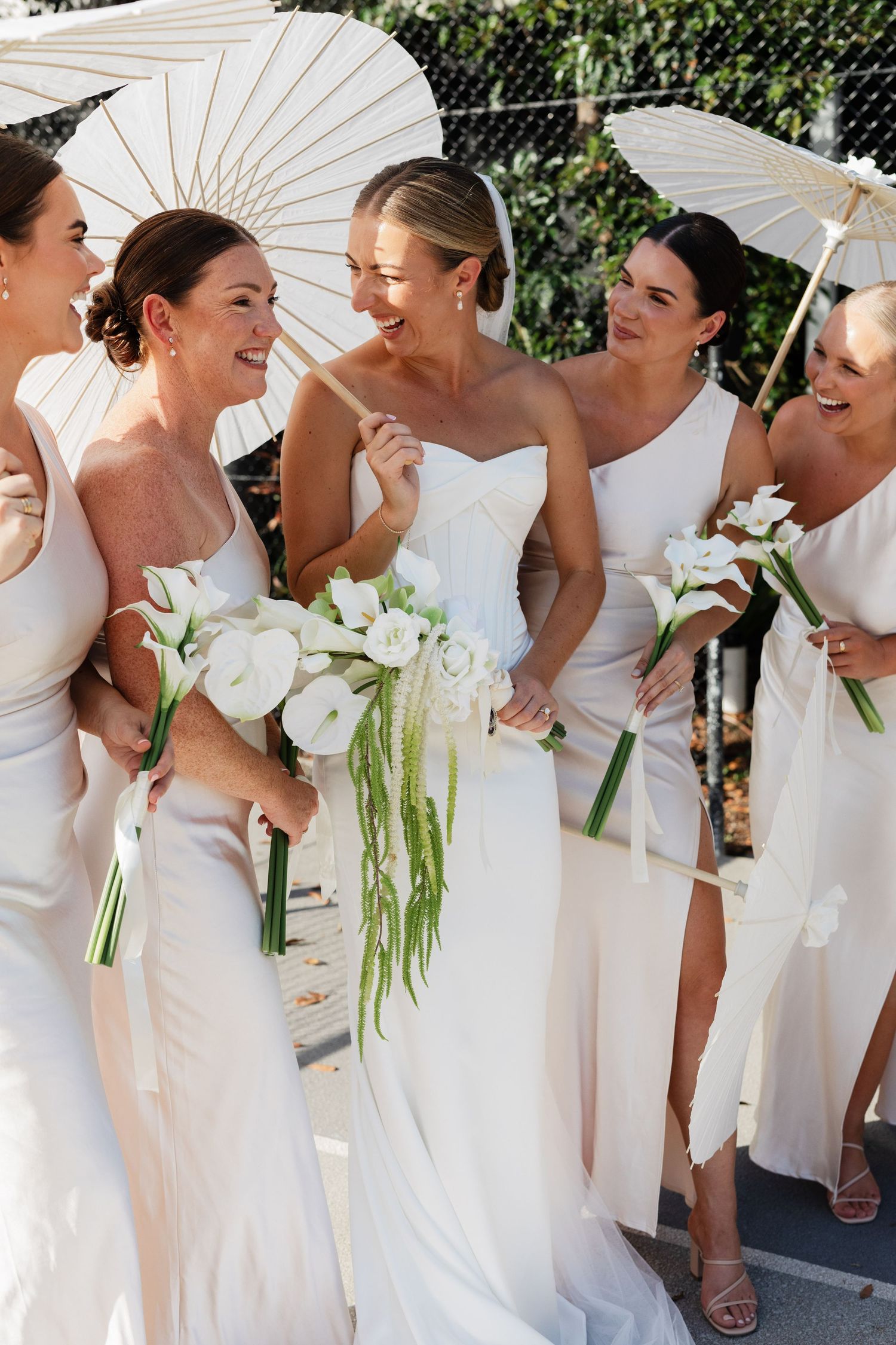 Bride in white gown laughs with bridesmaids in ivory dresses holding white floral bouquets and parasols outdoors.