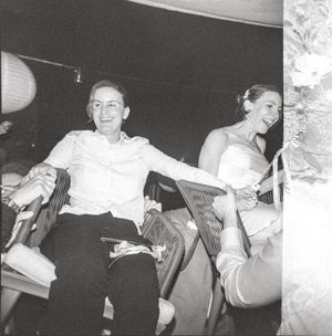 Two women laughing and holding hands at a wedding reception, one in a white dress, captured in black and white.