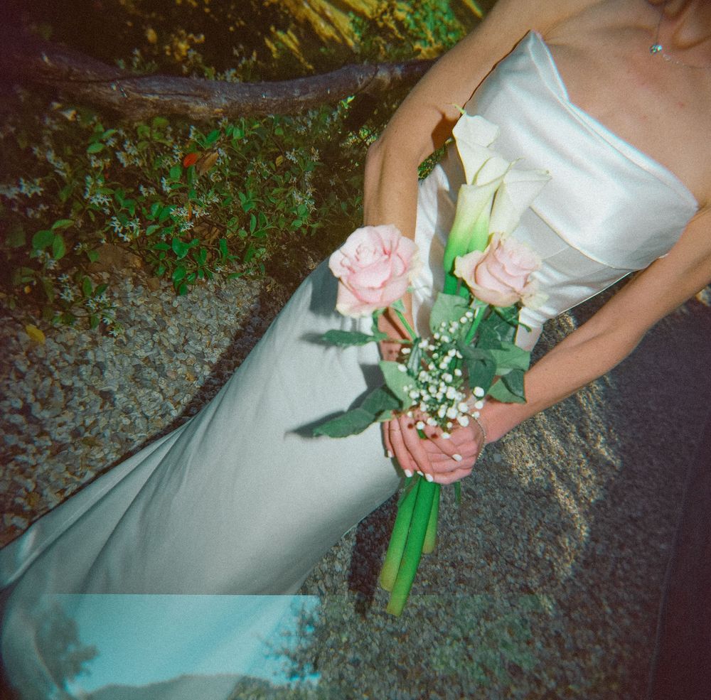 Bride in white strapless gown holding pink roses and calla lily bouquet with baby's breath at outdoor wedding.