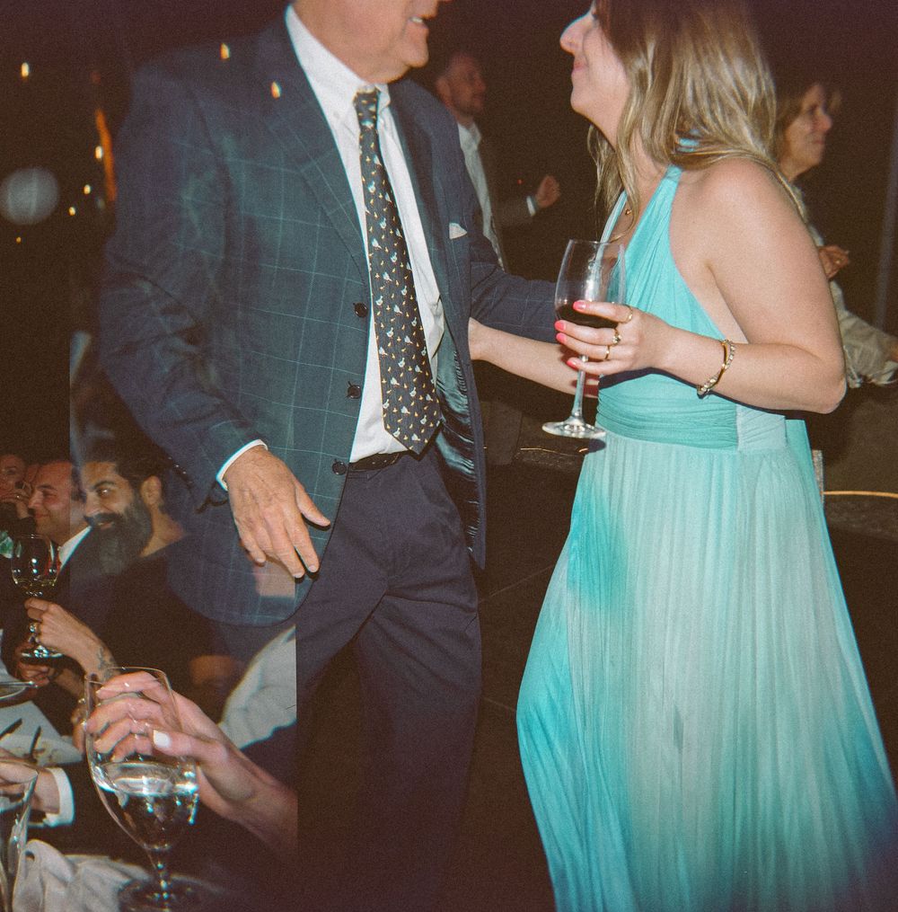 Man in blue plaid suit and woman in teal dress socializing at an elegant evening event with wine glasses.