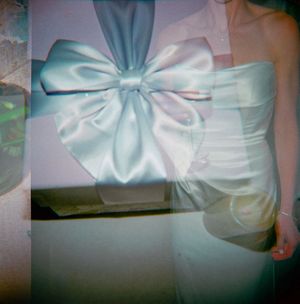 Elegant white satin gift box with large decorative bow, double-exposed with a woman in a strapless white gown.