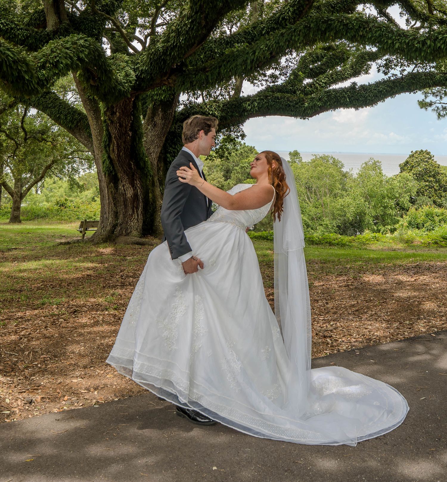 Bride and groom share romantic embrace under sprawling oak tree in lush green park setting.