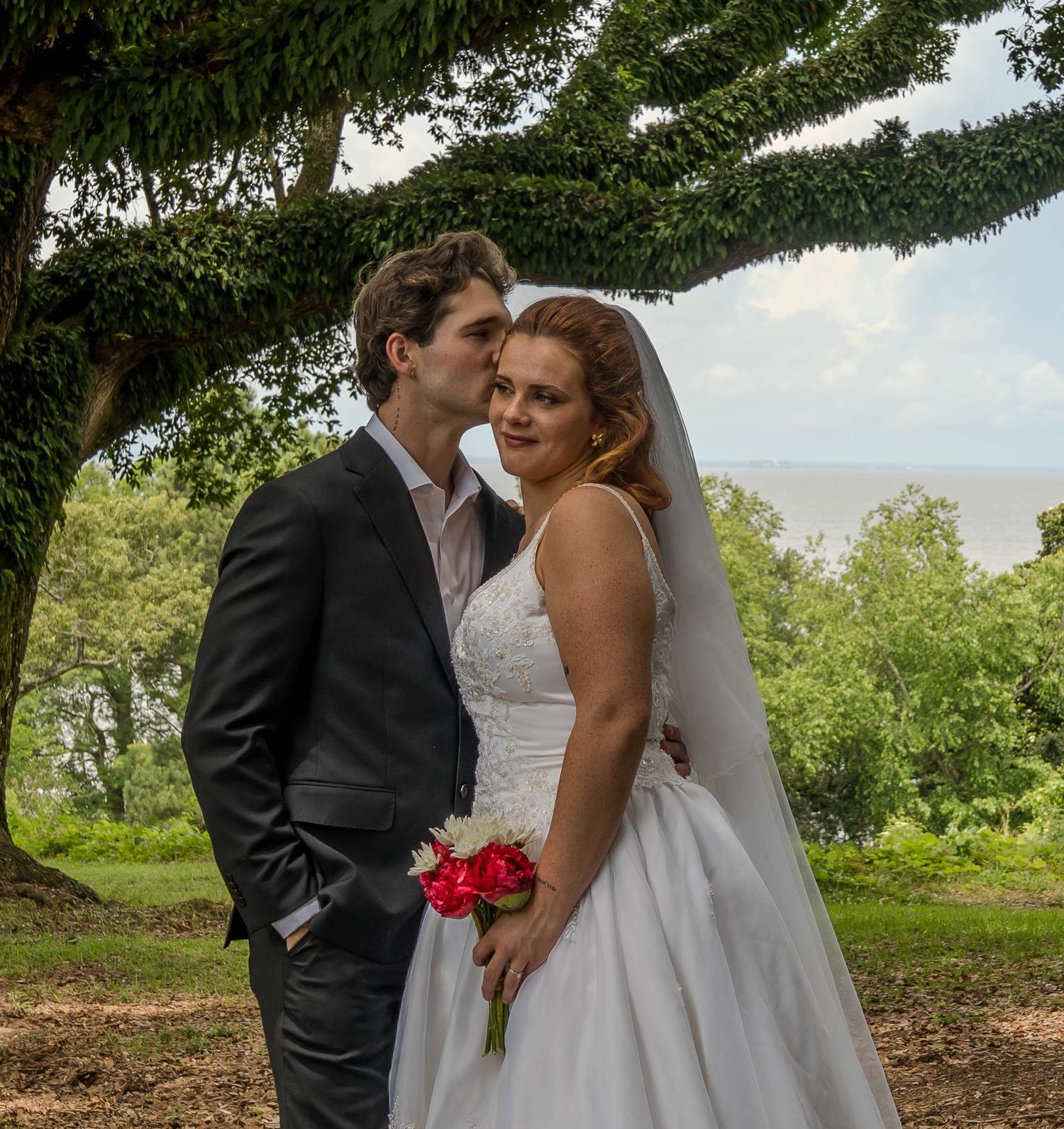 Bride and groom share a tender kiss under a lush tree outdoors, bride holding red roses in a white gown.