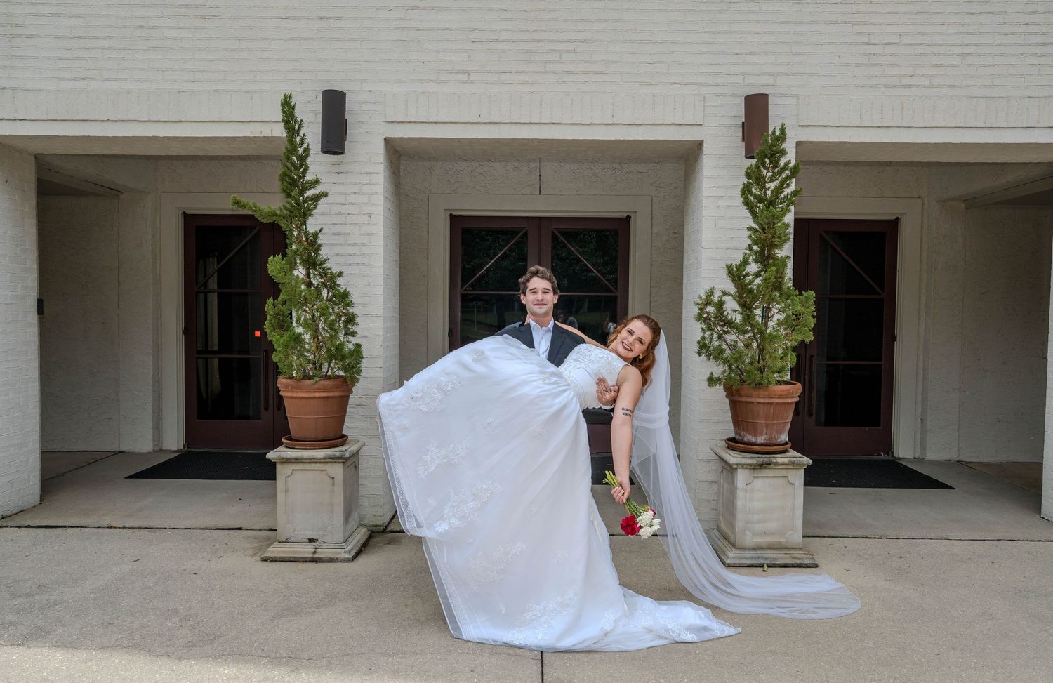 Groom lifting bride in white wedding dress outside elegant venue with potted plants and brick walls.