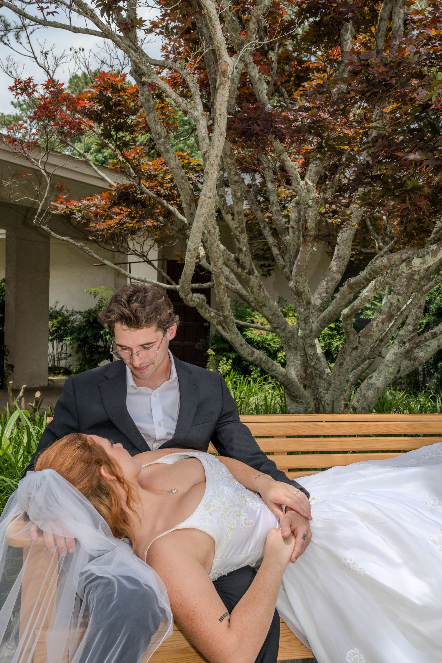 Groom sitting on a bench holding his bride in his arms, surrounded by colorful autumn trees.