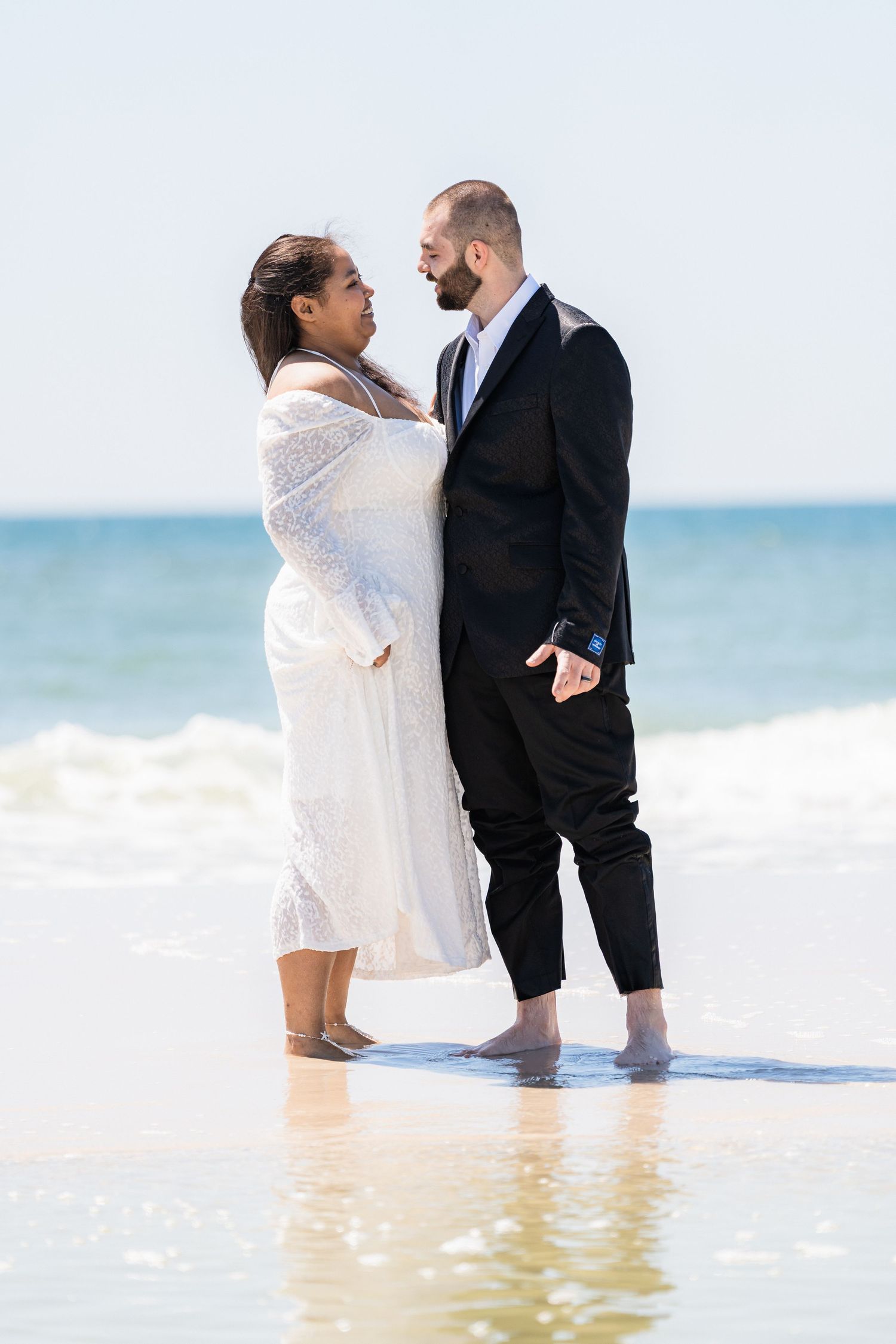 Couple stands barefoot in ocean waves, bride in white dress and groom in black suit, gazing at each other.