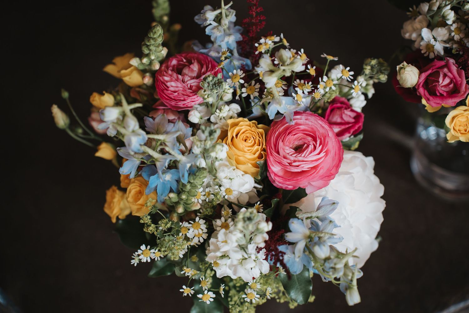 Colorful wedding bouquet featuring pink ranunculus, yellow roses, blue hydrangeas, and wildflowers on dark background.