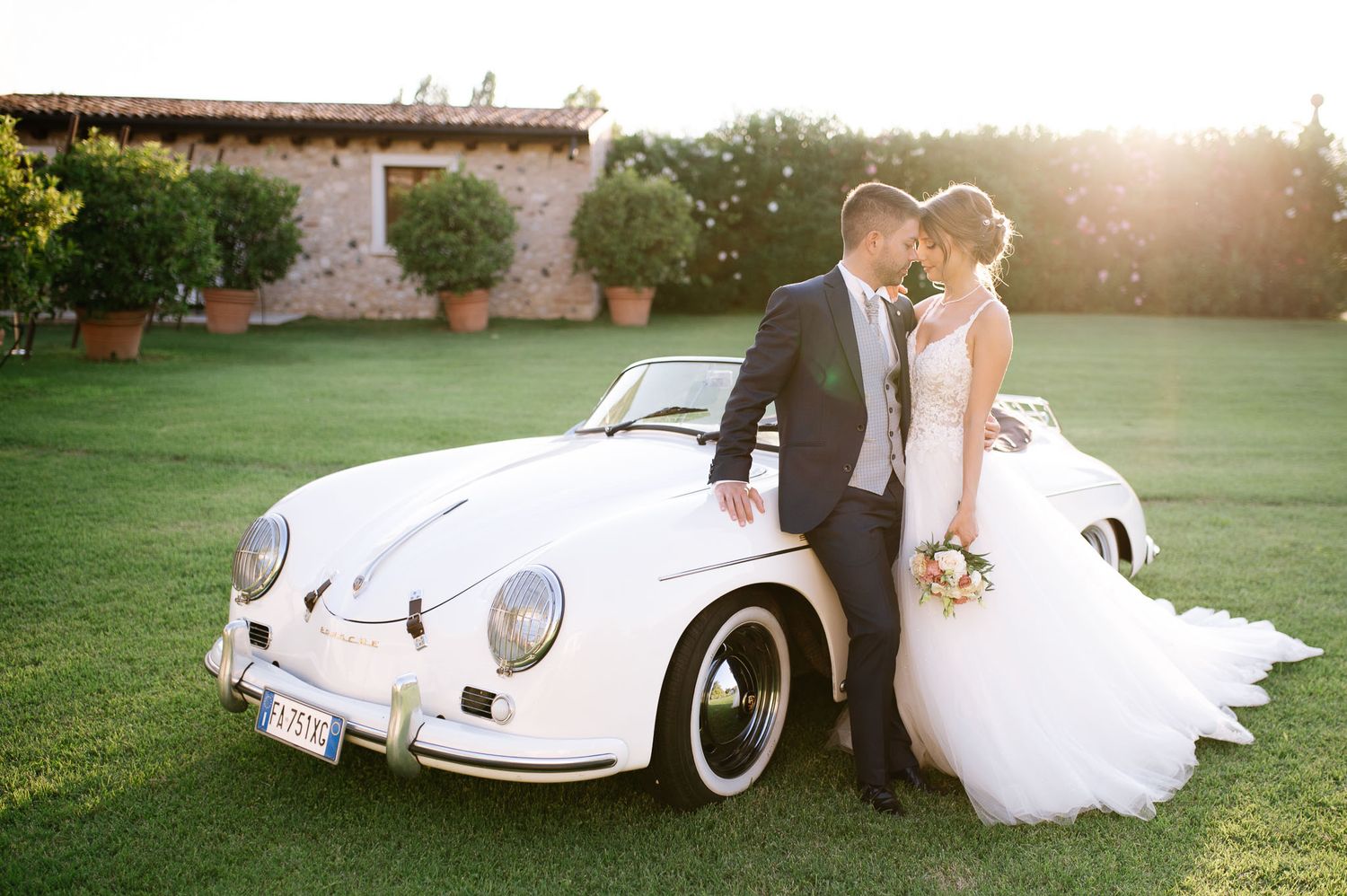 Bride and groom embracing beside a classic white Porsche 356 on a sunny lawn at a rustic Italian wedding venue.