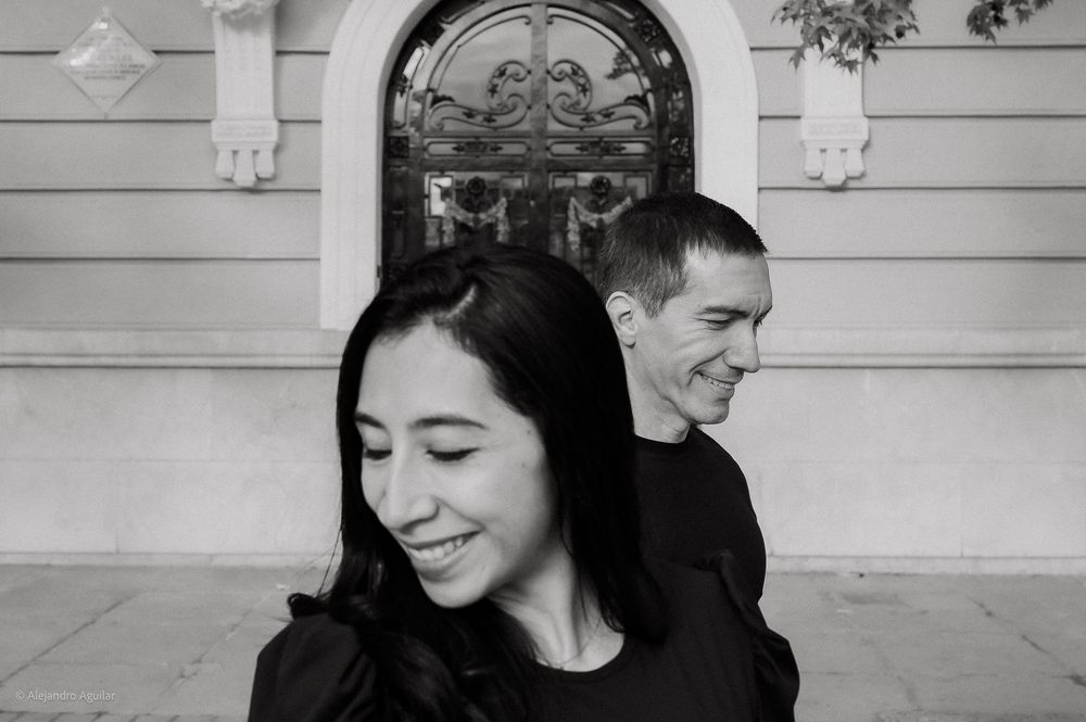 Black and white photo of smiling couple standing back to back before ornate building door.