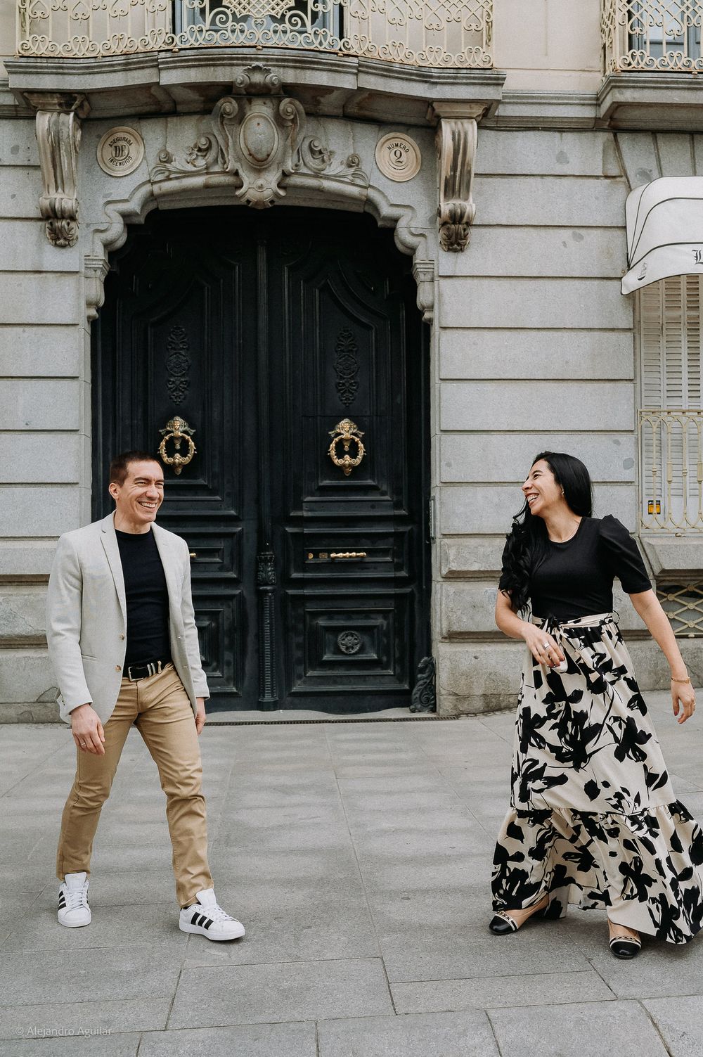 Happy couple laughing and walking together in front of ornate black doors of historic building.