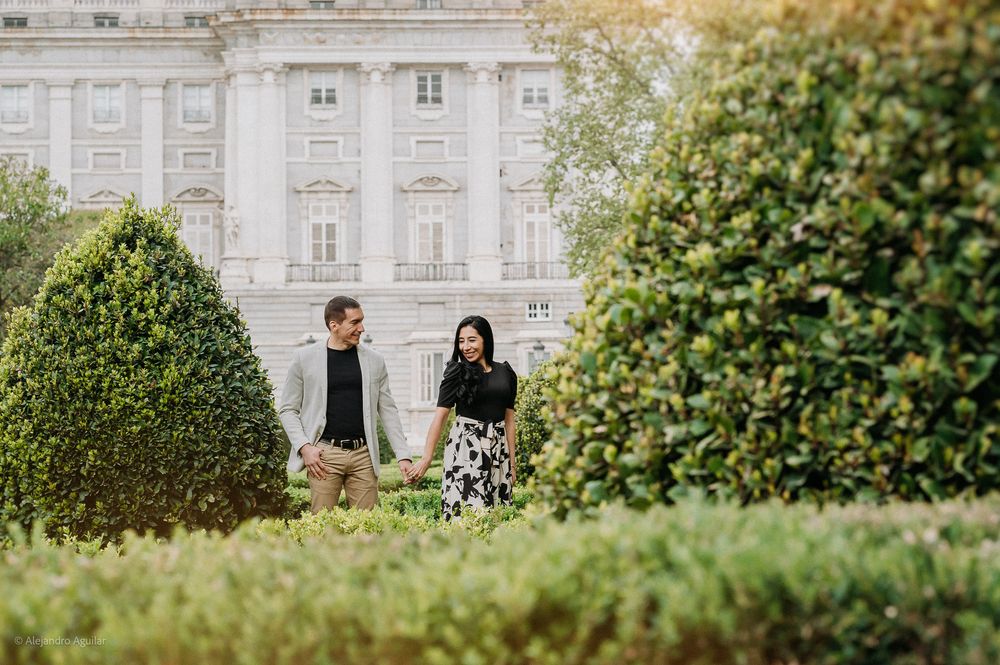Couple holding hands strolling through manicured garden with grand white palace in background.