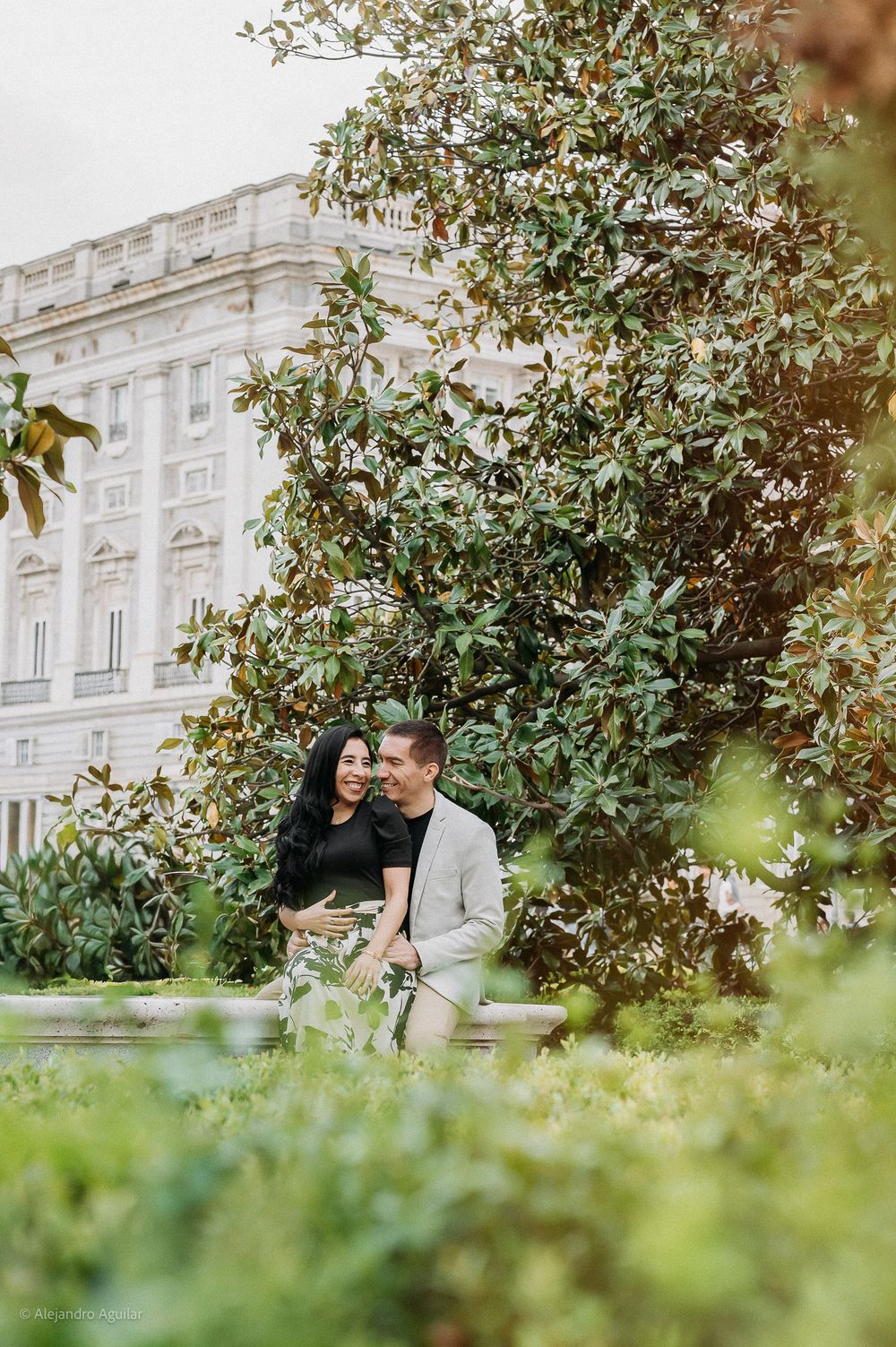 Couple sitting closely together on bench surrounded by lush greenery near white palace building.