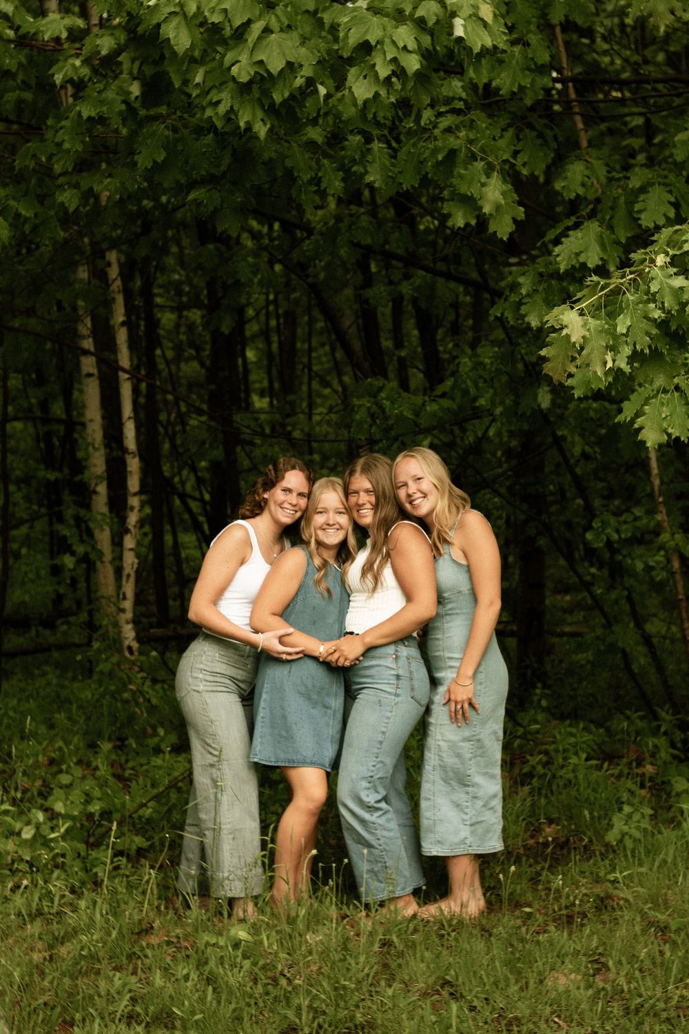Four smiling women in denim outfits pose together in a lush green forest setting.