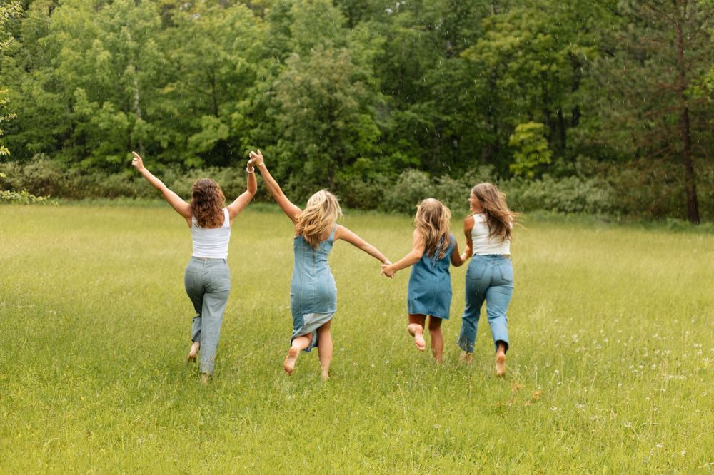 Four girls holding hands running through a green meadow with trees in the background on a sunny day.