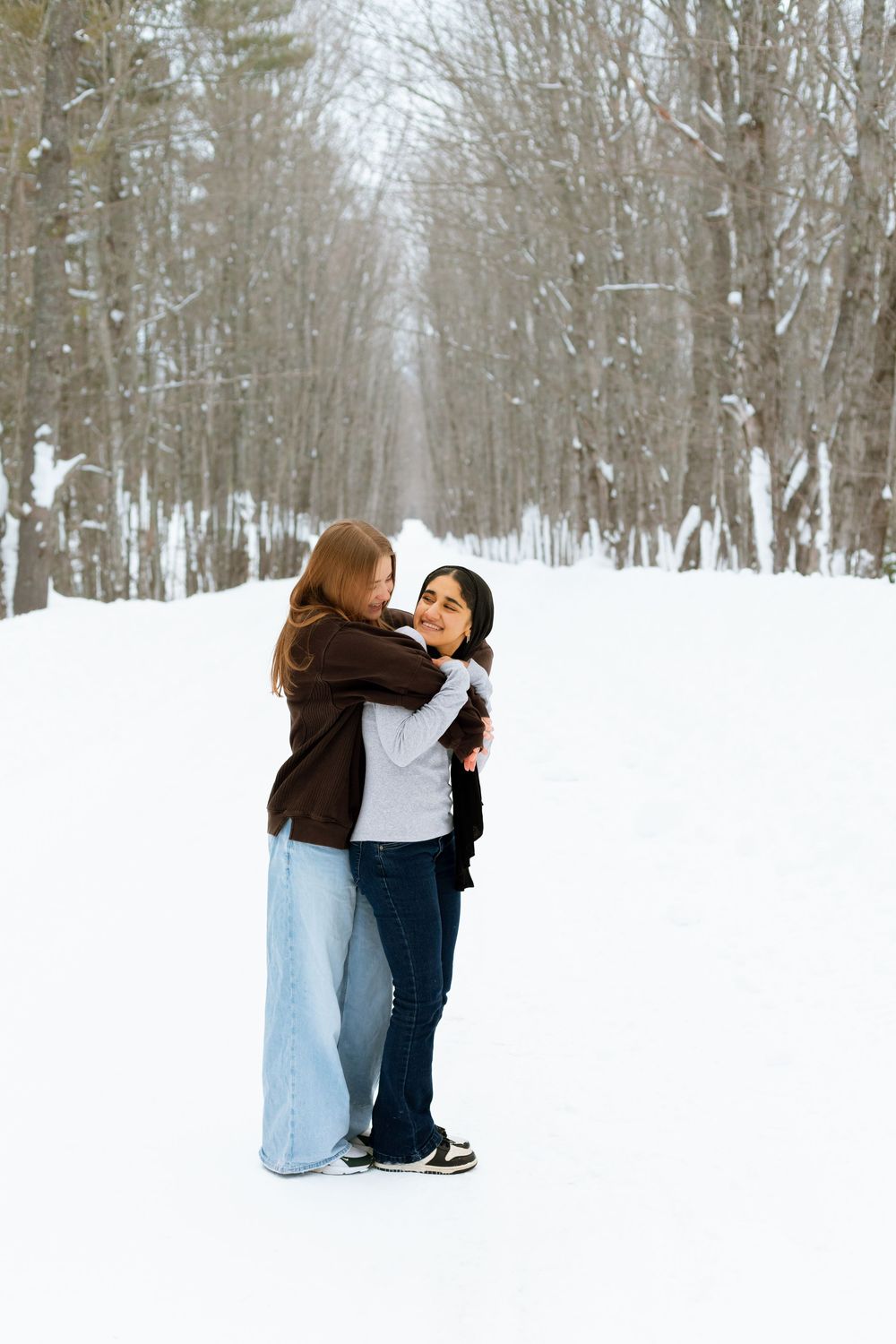Two women embrace warmly on a snowy winter road lined with bare trees.