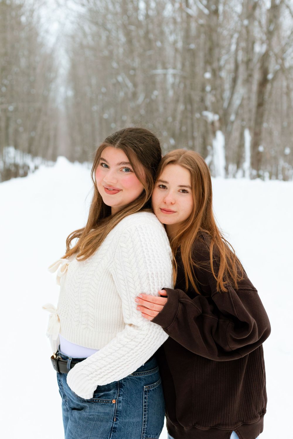 Two teenage girls in cozy sweaters embrace back-to-back in a snowy winter landscape surrounded by bare trees.