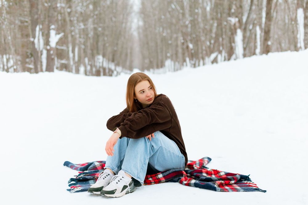 Young woman sitting on a plaid blanket in a snowy winter forest wearing jeans and a brown sweater with ice skates.