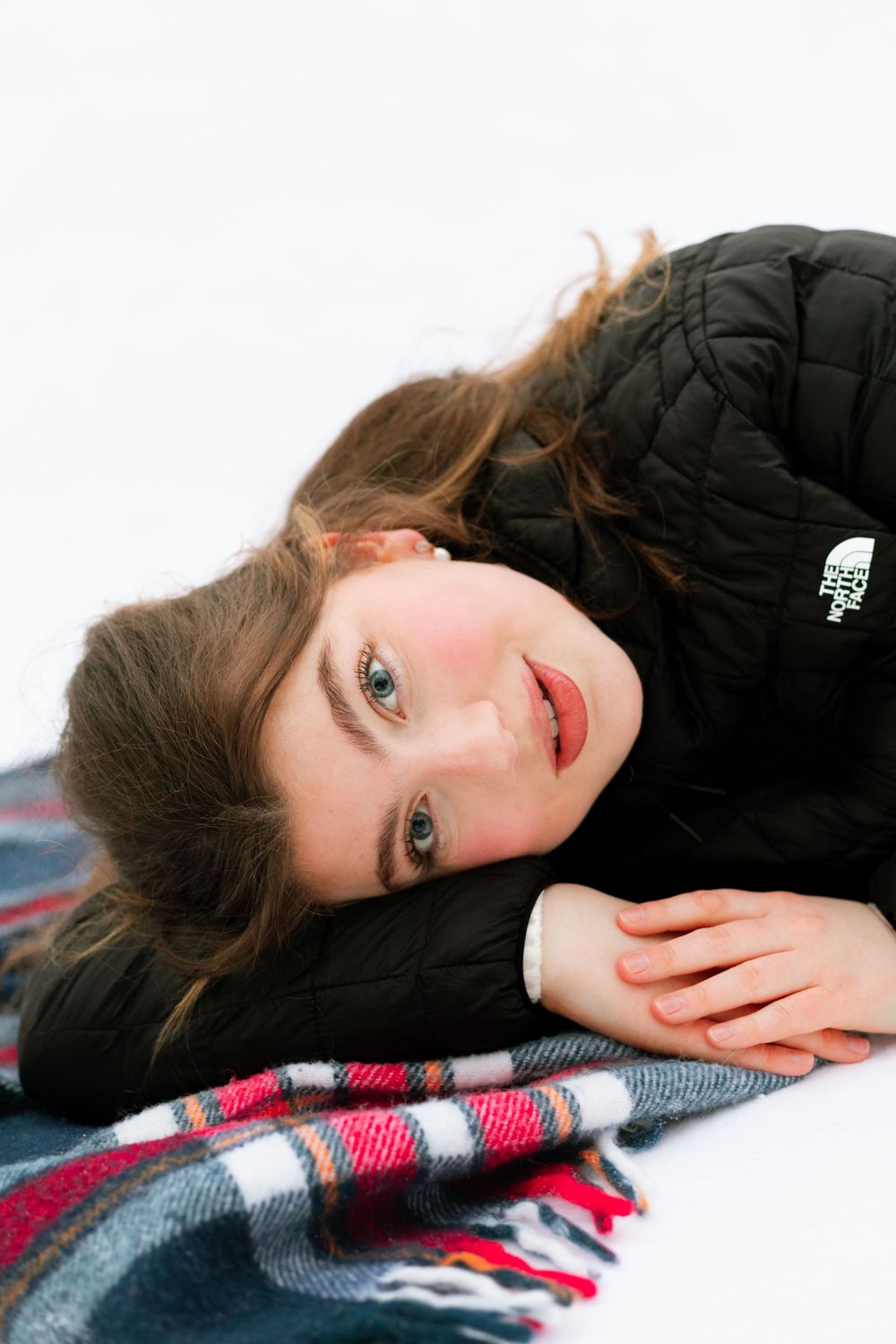 Young girl with blue eyes lying on plaid scarf in snow wearing black North Face jacket.