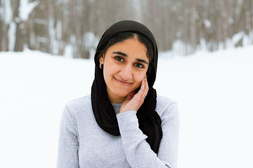 Young woman wearing a black hijab smiles outdoors in a snowy winter landscape, resting her hand on her cheek.
