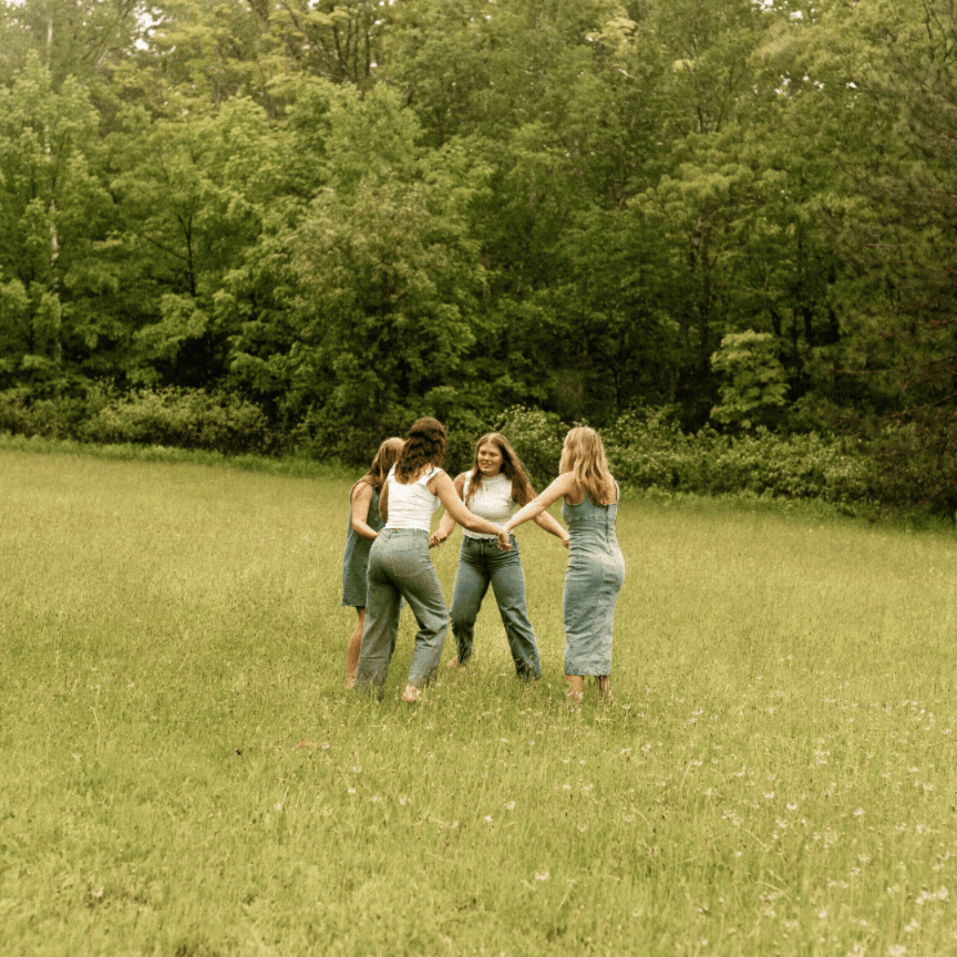 Four young women holding hands in a circle in a green meadow with lush forest in the background.